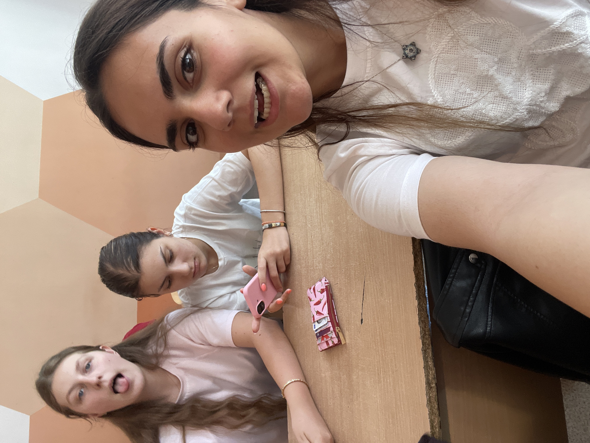Three young women are taking a selfie indoors. One woman is closest to the camera and smiling. A second woman is in the middle of the frame with her eyes closed. The third woman is at the bottom of the frame, sticking her tongue out. They are at a desk with two pink phones, a pink wallet, and a black jacket. The walls are painted with alternating diagonal sections of beige and peach. The location is Burgas, Bulgaria.