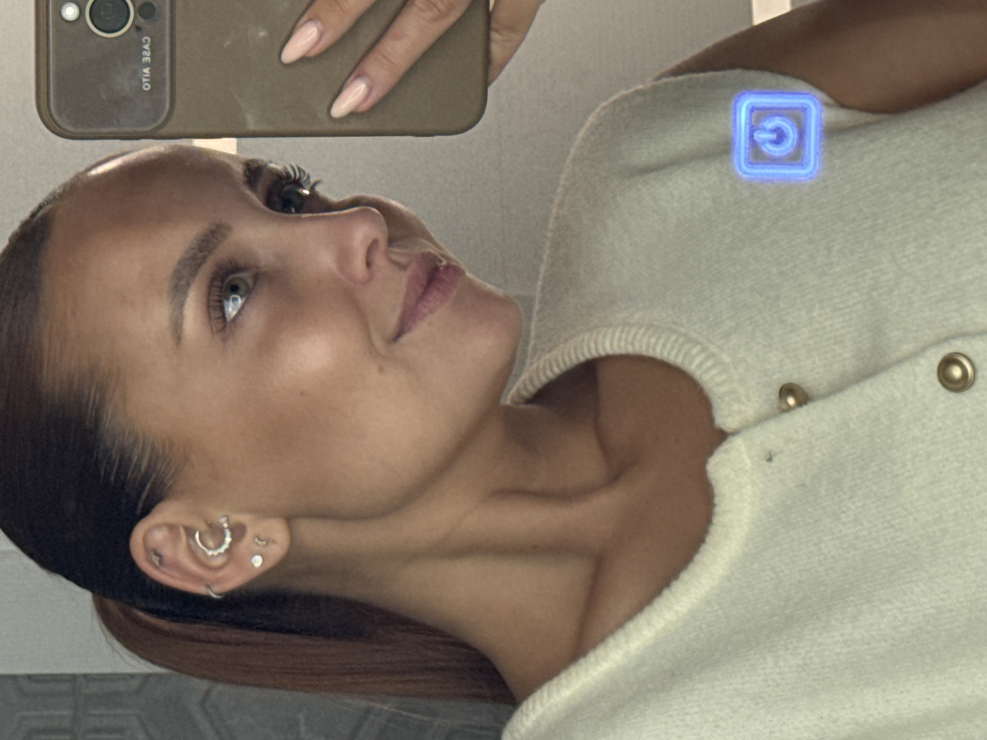 A young adult female with a sleek, dark brown ponytail and tanned skin is captured in a mirror selfie, seen in profile from the chest up. She has light-colored eyes, well-defined eyebrows, and a subtle smile. Her visible left ear features multiple piercings, including a prominent bejeweled hoop in the conch, a small stud and a tiny crescent moon stud in the lobe, and several other small studs or hoops along the helix. She wears an off-white or cream-colored, finely knit, sleeveless top or cardigan, secured with two visible round gold-toned buttons down the front. Her right hand, with almond-shaped nails painted in a light, neutral polish, holds a smartphone with a plain olive green or beige case. The phone's camera module is visible, and reversed text that appears to read "OTIA 32AJ" is reflected on its case. A glowing blue digital power symbol icon is superimposed on the upper chest area of her top, over her left shoulder. The background, visible through the mirror reflection, is a neutral-toned indoor wall, possibly tiled or paneled, with subtle horizontal lines. The image contains no visual information to indicate a specific geographic location.