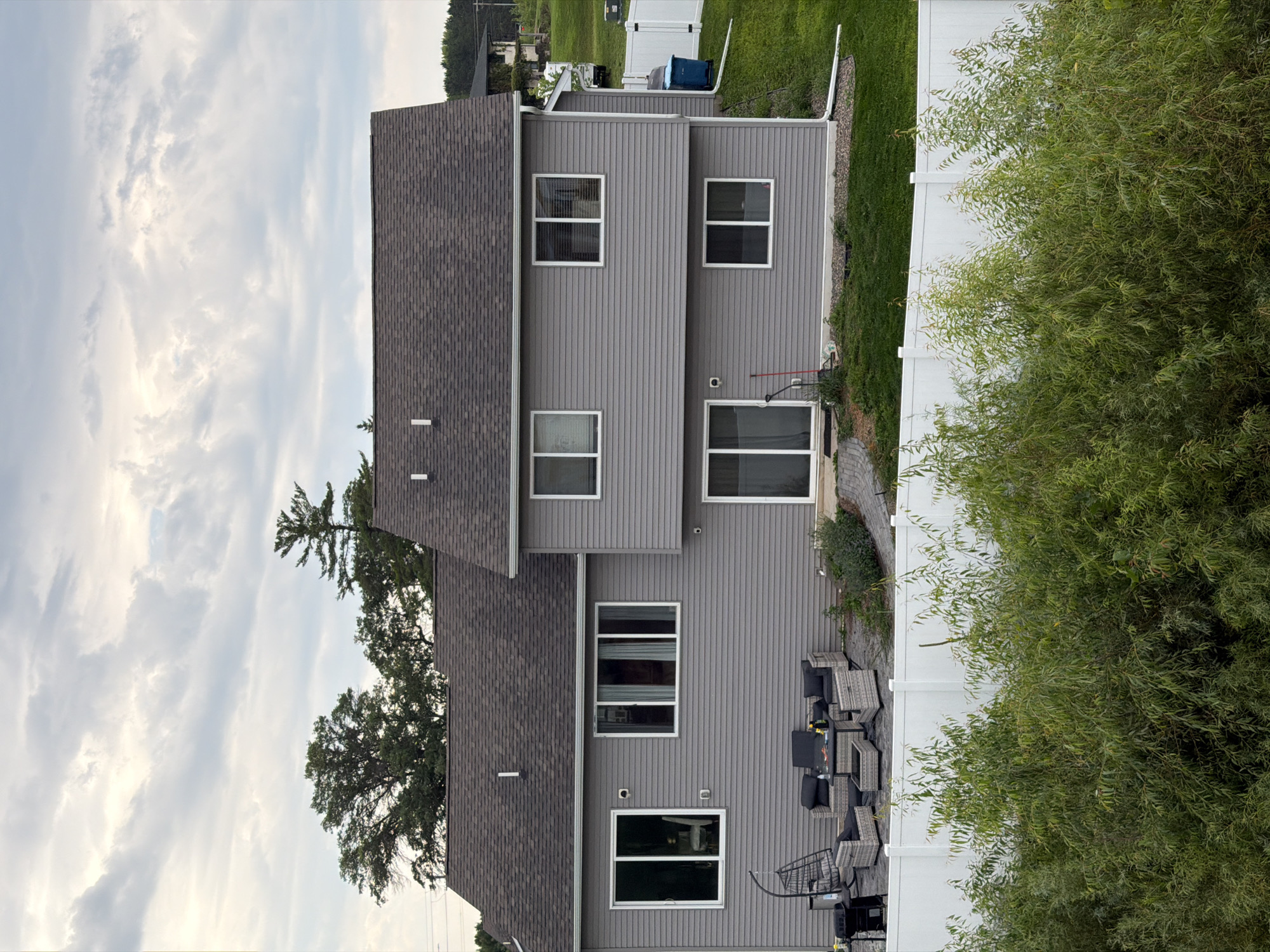 The image shows the rear exterior of a two-story gray vinyl-sided house with a dark gray asphalt shingle roof, located in Elk River, United States of America. The house has multiple windows, including a large sliding glass door on the lower level and several rectangular windows on both floors. In the backyard, there is a patio area with outdoor furniture, including a charcoal grill, a hanging chair, and a sectional sofa with a coffee table. A white vinyl fence runs along the perimeter of the yard, behind which are green lawn and some trees. A blue trash bin is visible next to the house near a white downspout. The sky is cloudy with scattered sunlight.