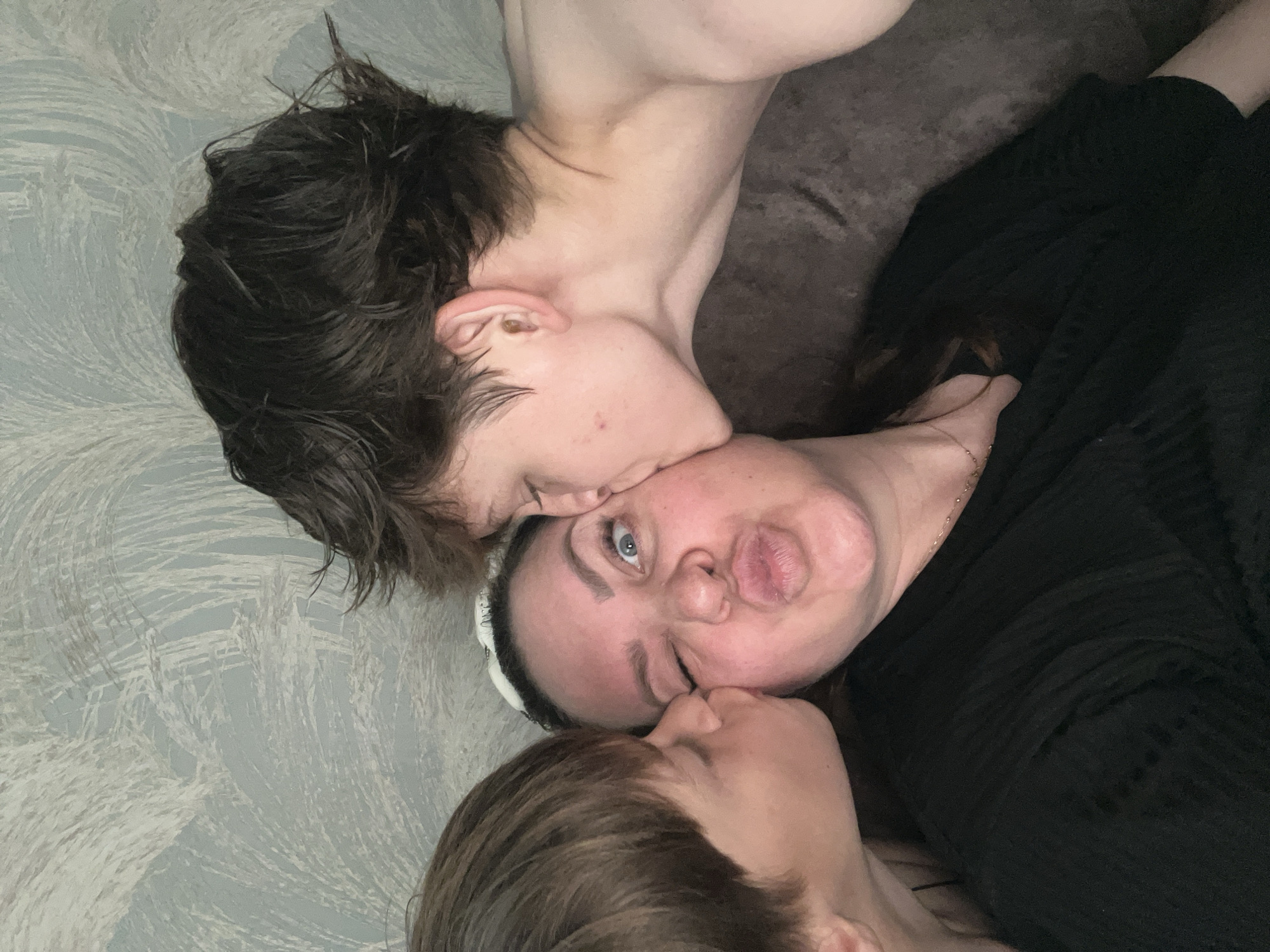 A close-up image captures three individuals in an indoor residential setting in the city of Noviy, Russia. A light-skinned adult female, centered in the frame, is being kissed simultaneously on both cheeks by two younger males. She wears a black short-sleeved top and a delicate gold-colored necklace. Her dark hair is pulled back, revealing a white headband. Her left eye is closed in a wink, and her lips are puckered outward in a playful gesture, while her right eye is wide open, revealing a light blue iris. The younger male on the left, with short brown hair, is shown in profile kissing her left cheek; only his head and ear are visible. The second younger male on the right, with darker, messy hair, is also shown in profile, kissing her right cheek; he appears shirtless, with a bare shoulder and neck visible, and a small red blemish is present on his right cheek. The background consists of light blue/grey wallpaper with a subtle, swirling white abstract pattern on the upper left, and dark grey/brown textured upholstery, likely a couch or cushion, on the lower right. The interaction conveys a moment of affectionate familial playfulness.