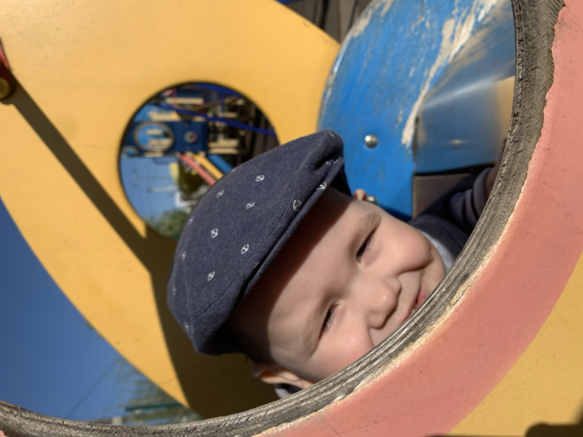 A close-up shows a child wearing a blue newsboy cap with small, light-colored anchor patterns, peeking through a circular opening of a playground structure. The structure is painted in yellow, blue, and pinkish-red. The child has fair skin and a slight smile. The background reveals other parts of the playground structure and a glimpse of blue sky. The location is Фоки, Russia.