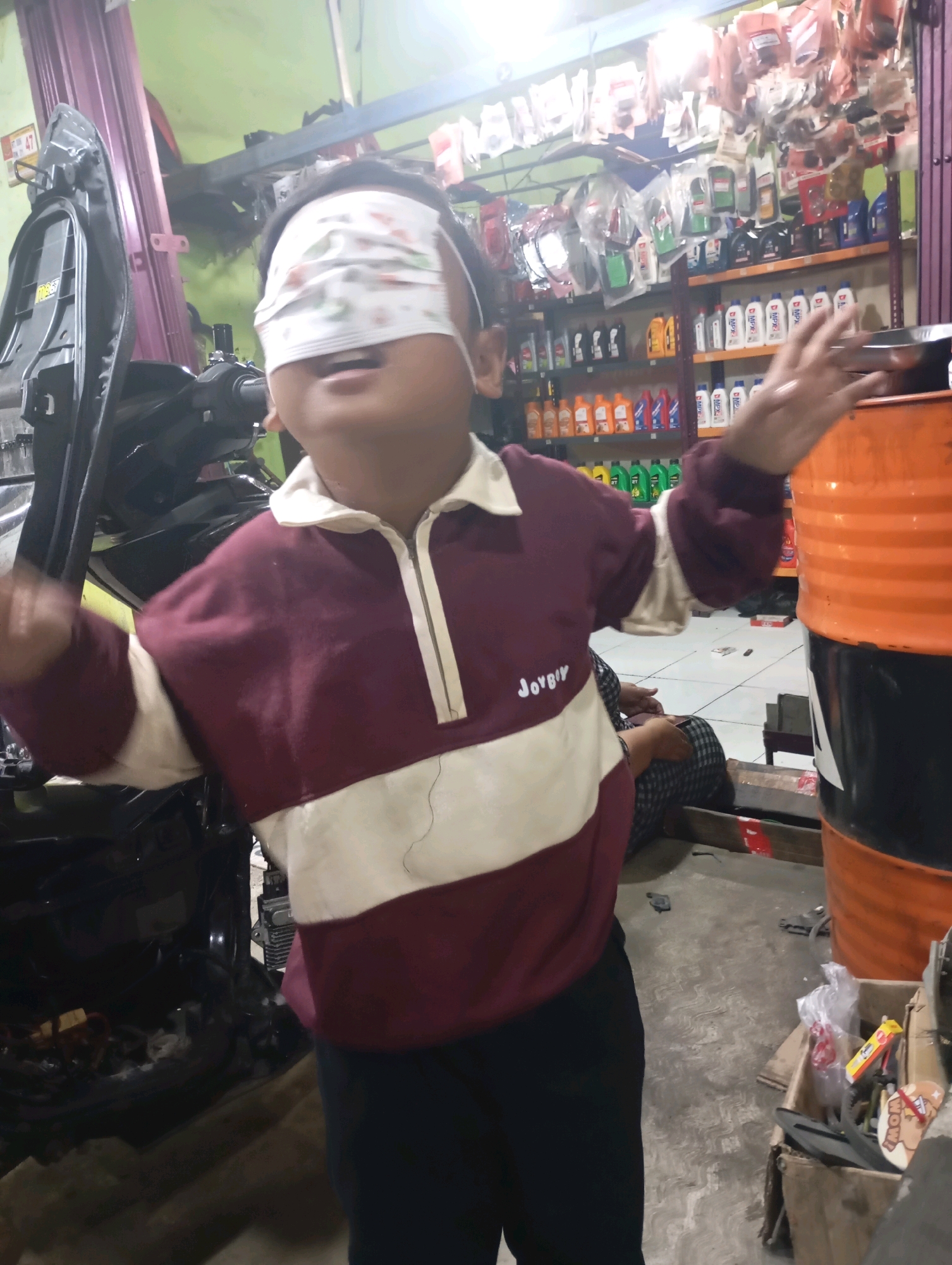 In a retail setting resembling a motorcycle parts shop in Babelan, Indonesia, a young individual is depicted with a fabric mask covering their eyes, obscuring their vision. The individual is wearing a maroon and cream-colored, zippered sweatshirt. To the left, a motorcycle is partially visible. The background features shelves stocked with various automotive fluids, likely oil and lubricants, and other assorted items. An orange industrial drum is situated to the right of the individual, and a wooden crate containing miscellaneous items is in the foreground. The overall environment suggests a commercial establishment.