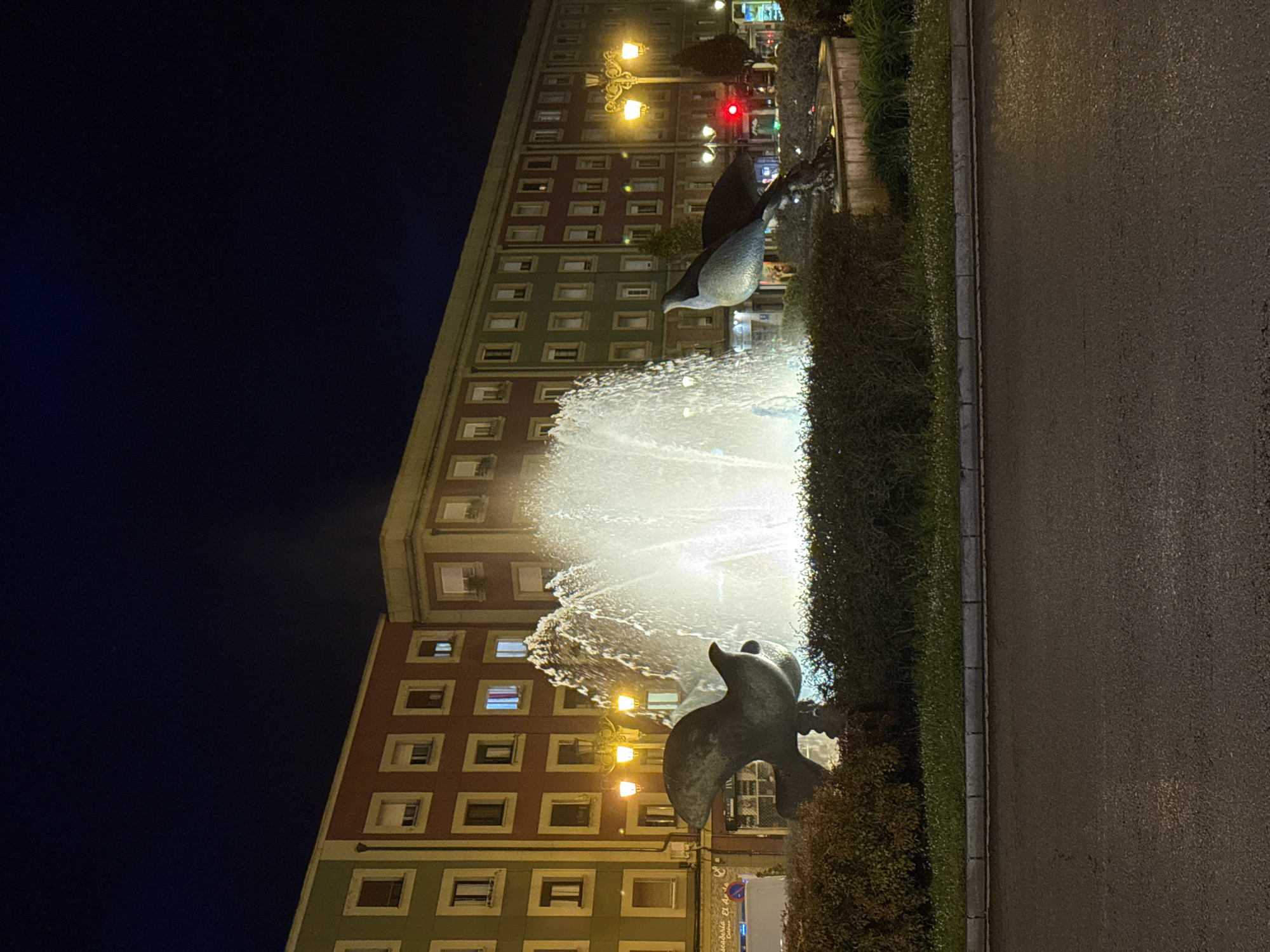 The image depicts the Uria Street fountain, a notable landmark in Oviedo, Spain, during nighttime. The fountain features bronze statues of dolphins, with water jets illuminating the scene. In the background, a large building with multiple windows, displaying a combination of red, green, and beige colors, is visible. Streetlights cast a warm glow, adding to the urban ambiance. A sidewalk and a street are observed on the right side of the frame.