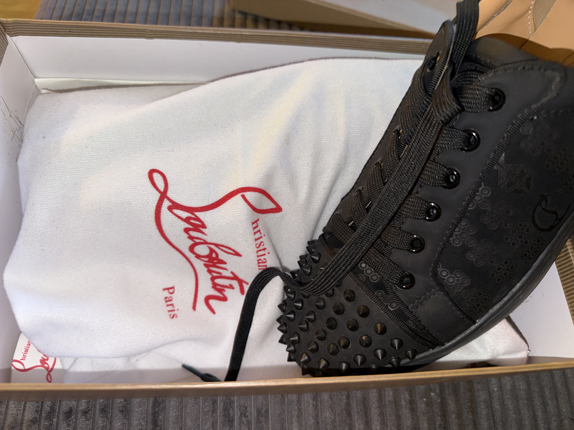 A black Christian Louboutin sneaker with black spikes and laces rests in an open shoebox. A white dust bag with the red Christian Louboutin logo and "Paris" text is partially visible behind the shoe. The background is a textured grey surface. The scene is indoors, and no specific location in Varna, Bulgaria, can be identified.