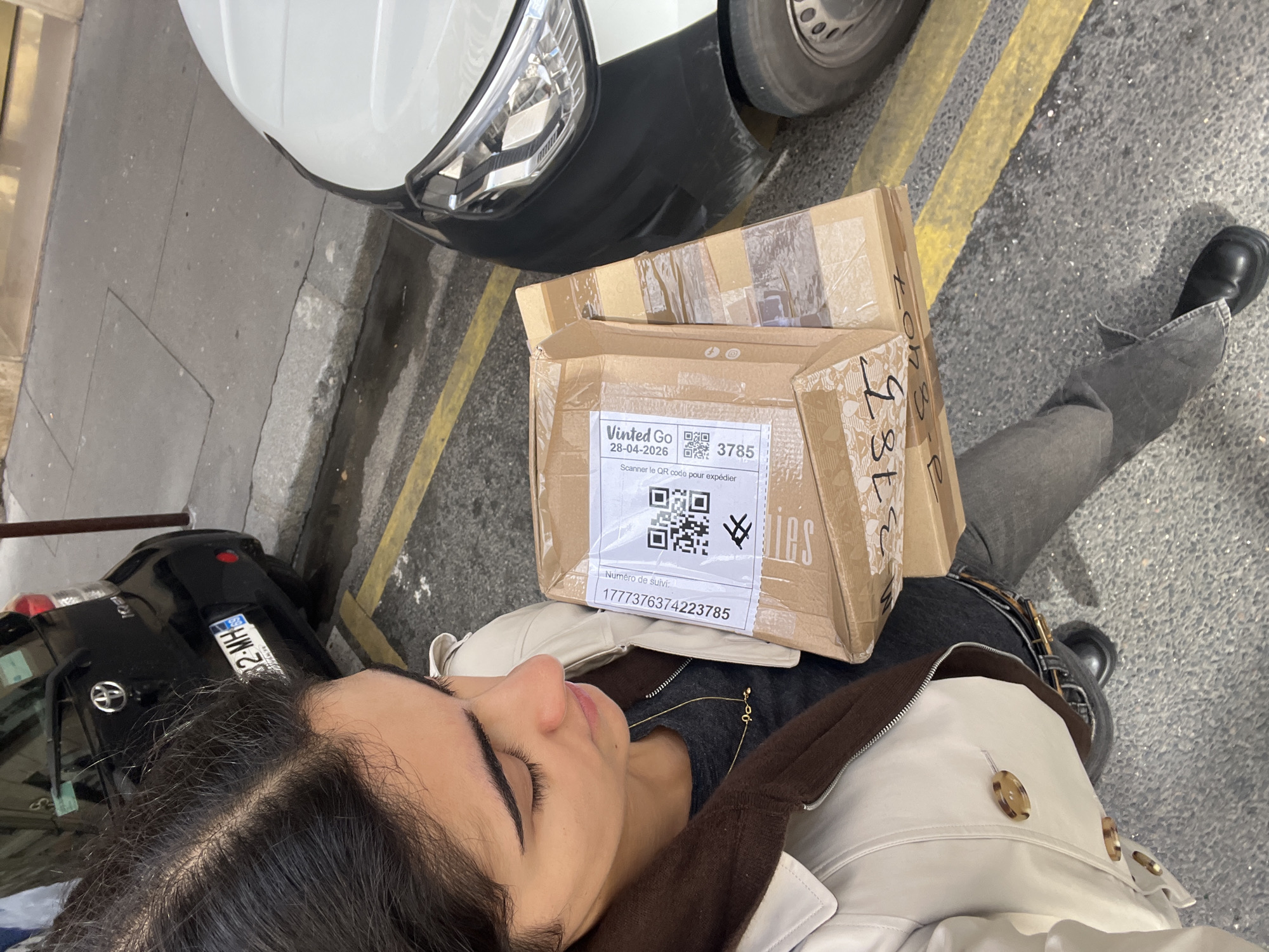 From a high-angle perspective, the image shows a woman standing on a street in Paris, France. She is wearing a light-colored jacket with brown accents, dark jeans and black boots. She is holding a brown cardboard package, marked with "Vinted Go" and a tracking number "1777376374223785," and bearing a QR code for scanning. The package appears to be resting against her. The edge of a black car and a white van are visible on the left and top of the frame. The street is marked with parallel yellow lines. A license plate is visible on the black car.