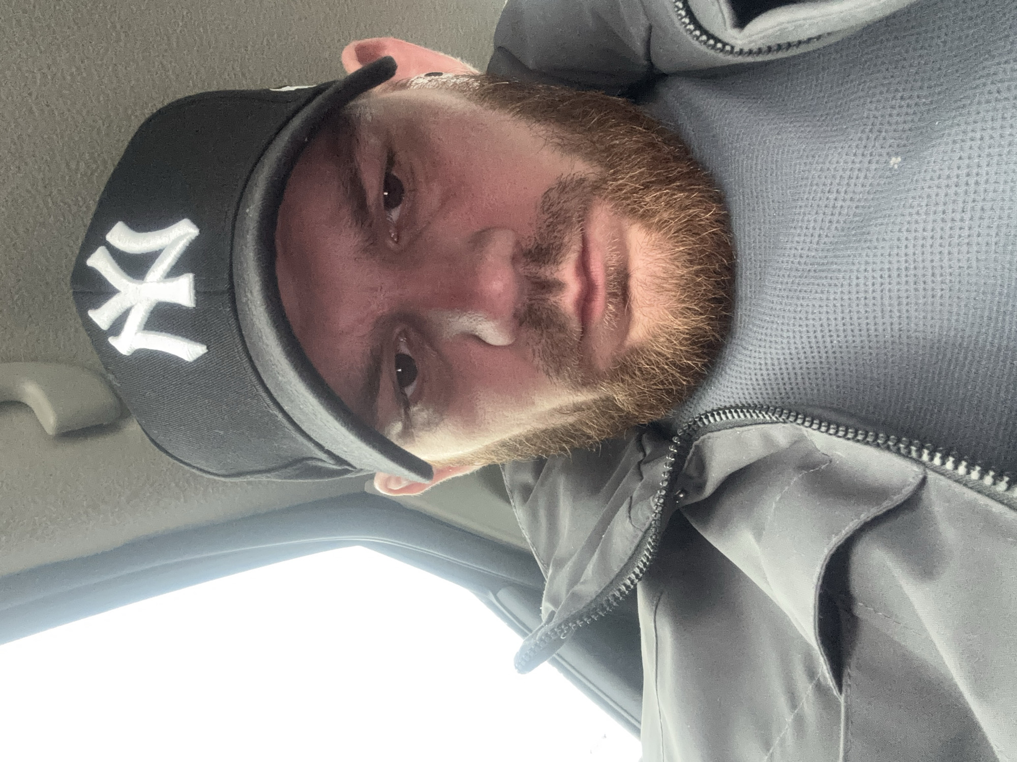 The image captures a close-up, low-angle portrait of a man inside a vehicle, located in Perm, Russia. The subject is a man in his 20s or 30s, with a reddish-brown beard and mustache, and dark eyes. He has a neutral to slightly serious expression as he looks directly into the camera.
He is wearing a black baseball cap with a prominent white "NY" logo, typically associated with the New York Yankees. His attire includes a dark grey or black zippered jacket layered over a lighter grey waffle-knit long-sleeved shirt.
The setting appears to be the interior of a car or van, with a light-colored headliner visible above him. A bright, diffused light source, consistent with an overcast sky seen through a vehicle window or windshield, illuminates the scene from below, indicating it is daytime. The image is either a selfie or a very close-up portrait, capturing the man from a slightly upward angle. The visible text is the "NY" logo on his cap.