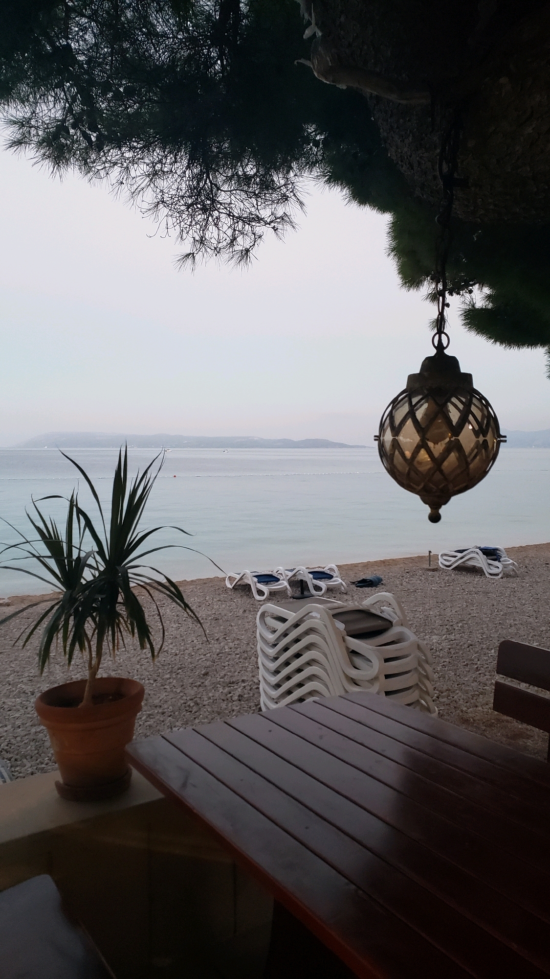 This is an outdoor scene at a beachside establishment, likely in Općina Tučepi, Croatia, given the context of similar images. In the foreground, a dark wooden table with visible planks is positioned centrally. To the left, a terracotta pot contains a spiky green plant, sitting on a low concrete structure. Behind the table and plant, the beach is covered in small gray pebbles. Several stacks of white plastic lounge chairs are arranged on the beach.
The background reveals a calm, pale blue sea under an overcast sky. In the distance, a faint coastline with hills is visible. A decorative, glass and metal lantern hangs from above on the right side of the frame, partially obscuring the view of the sky and pine tree branches. The overall atmosphere is serene and possibly twilight or early morning, given the soft lighting. There are no people visible in the image.