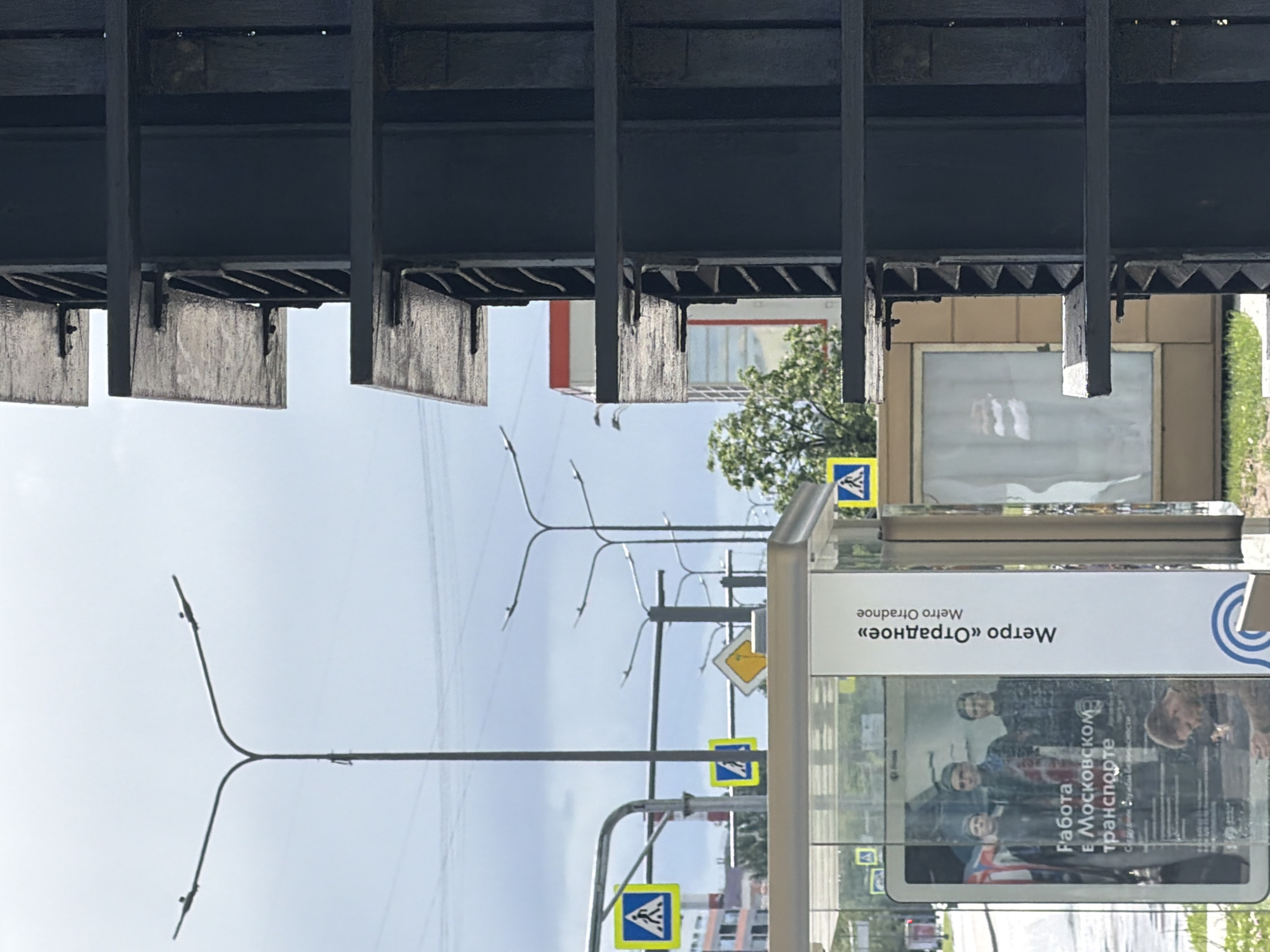 The image depicts an urban scene, possibly a street in the city of Moscow. The foreground features a bus stop with an advertising poster showcasing people and text in Russian. The poster mentions "Metro Otradnoe" and "Work in Moscow transport". Pedestrian crossing signs are visible. In the background, there are lamp posts, power lines, buildings, and foliage. A section of a metallic overhead structure is visible at the top of the frame. The environment is daytime under a cloudy sky.