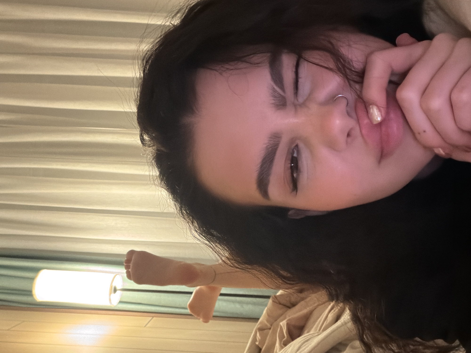 A close-up, slightly angled shot features a young woman indoors. She has dark hair, fair skin, and a nose ring. Her right eye is open and visible, while her left eye is squinted. She is holding her fingers to her lips, and her nails have a metallic polish. She appears to be wearing a dark-colored garment.
In the background, to the left, is a floor lamp with a cylindrical shade casting a warm glow. Behind the lamp are light-colored vertical blinds. There is a visible arm and closed fist near the bottom left of the frame, attached to a thin bracelet. The lower part of the image shows a beige-colored blanket or duvet.
The location is unknown. There is no information to define the city or environment.