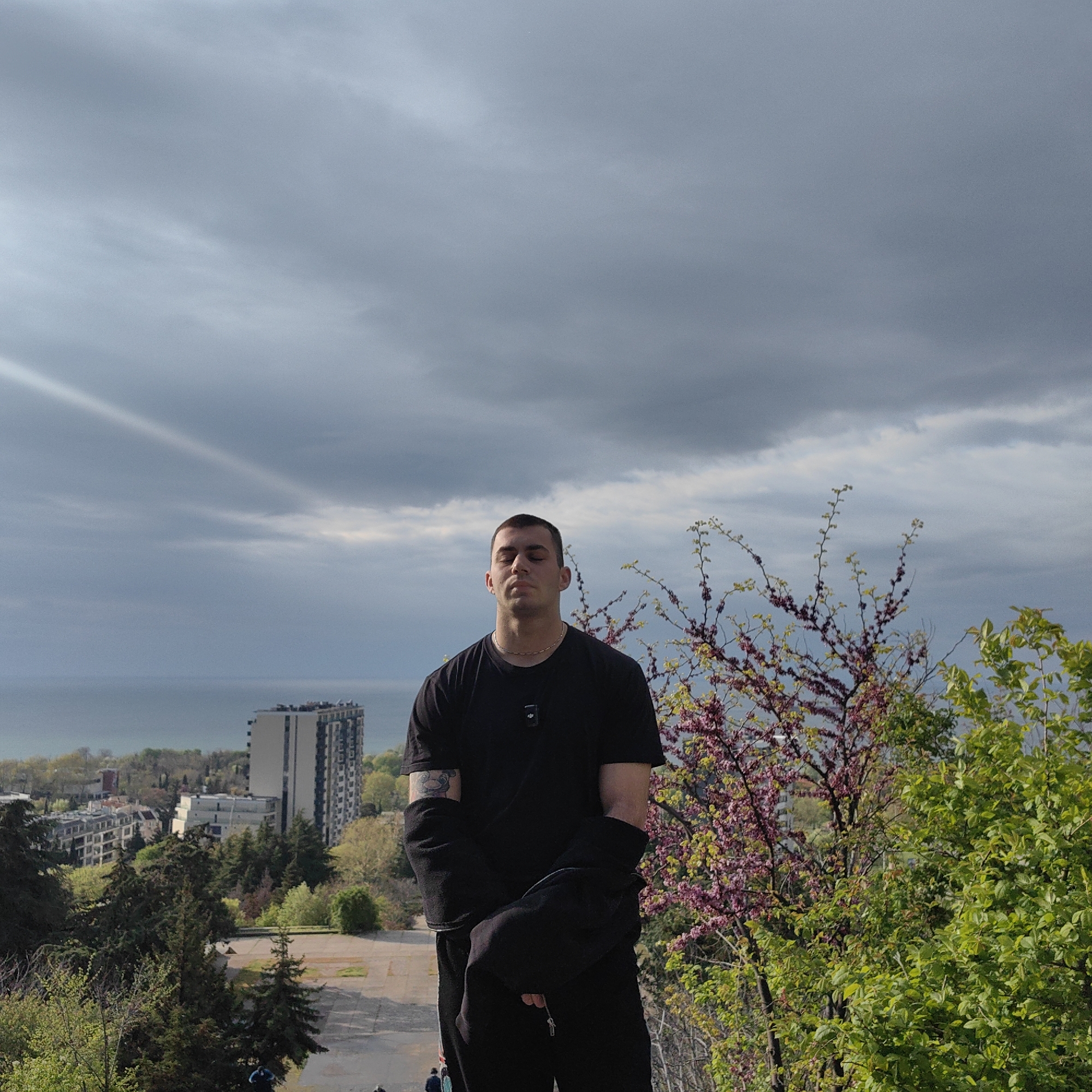 A young man with closely cropped dark hair, wearing a black t-shirt and dark pants, stands with his eyes closed. He is positioned in front of a cityscape and seascape. The building behind him is likely in Varna, Bulgaria. The trees behind the man have green foliage and some pink blossoms. The sky above is cloudy. A ray of light or rainbow segment is visible to the left of the frame in the sky. In the background, a tall, modern building stands among other buildings. A sea expanse is visible in the distance. A black zip-up jacket is slung over his arms.