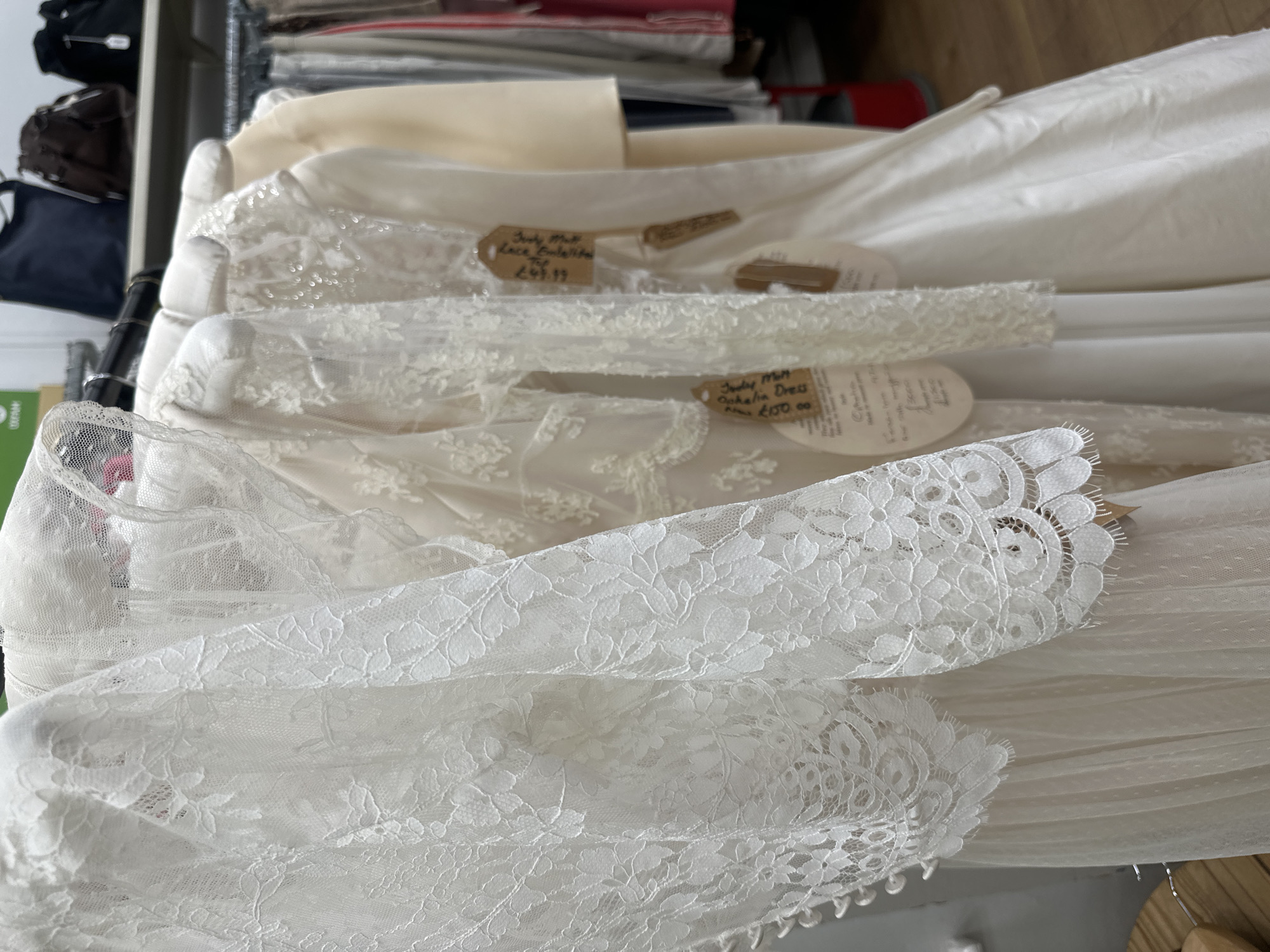 The image captures a close-up view of several white lace garments hanging on a rack, likely wedding dresses or bridal attire. A price tag on one of the dresses reads "Judy Mott," suggesting the clothing is from a designer or boutique with that name. The location is likely a retail space in Tunbridge Wells, United Kingdom, as indicated by the price tag. The garments are predominantly white and feature intricate lace detailing, with different patterns and textures visible. The focus is on the delicate lace and the overall aesthetic of bridal wear.
