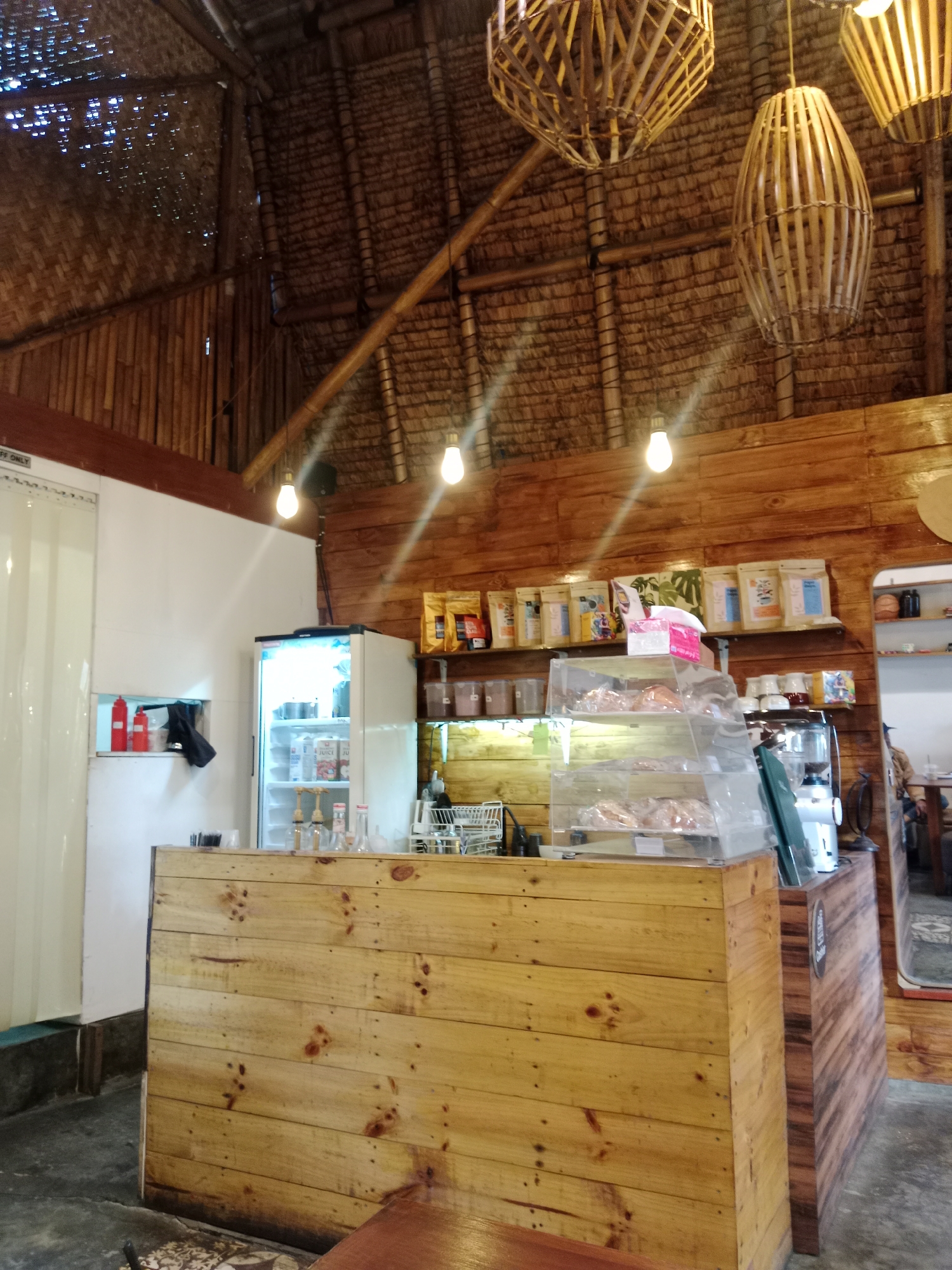 The image shows the interior of a cafe with a rustic design. The ceiling is made of bamboo and thatch, with three decorative pendant lights made of woven bamboo. The walls are paneled with wood. In the foreground, a wooden counter is visible, with a glass display case on top showcasing pastries. To the left of the counter, there is a white refrigerator with its door open, revealing shelves stocked with various items. Behind the counter, shelves are lined with bags of coffee or other products and containers. A coffee machine is visible on the right side of the counter. The floor is concrete, with a patterned rug visible in the lower left corner. The overall impression is one of a casual and cozy cafe environment. The image does not provide any specific information to suggest a location in Bandung City, Indonesia.