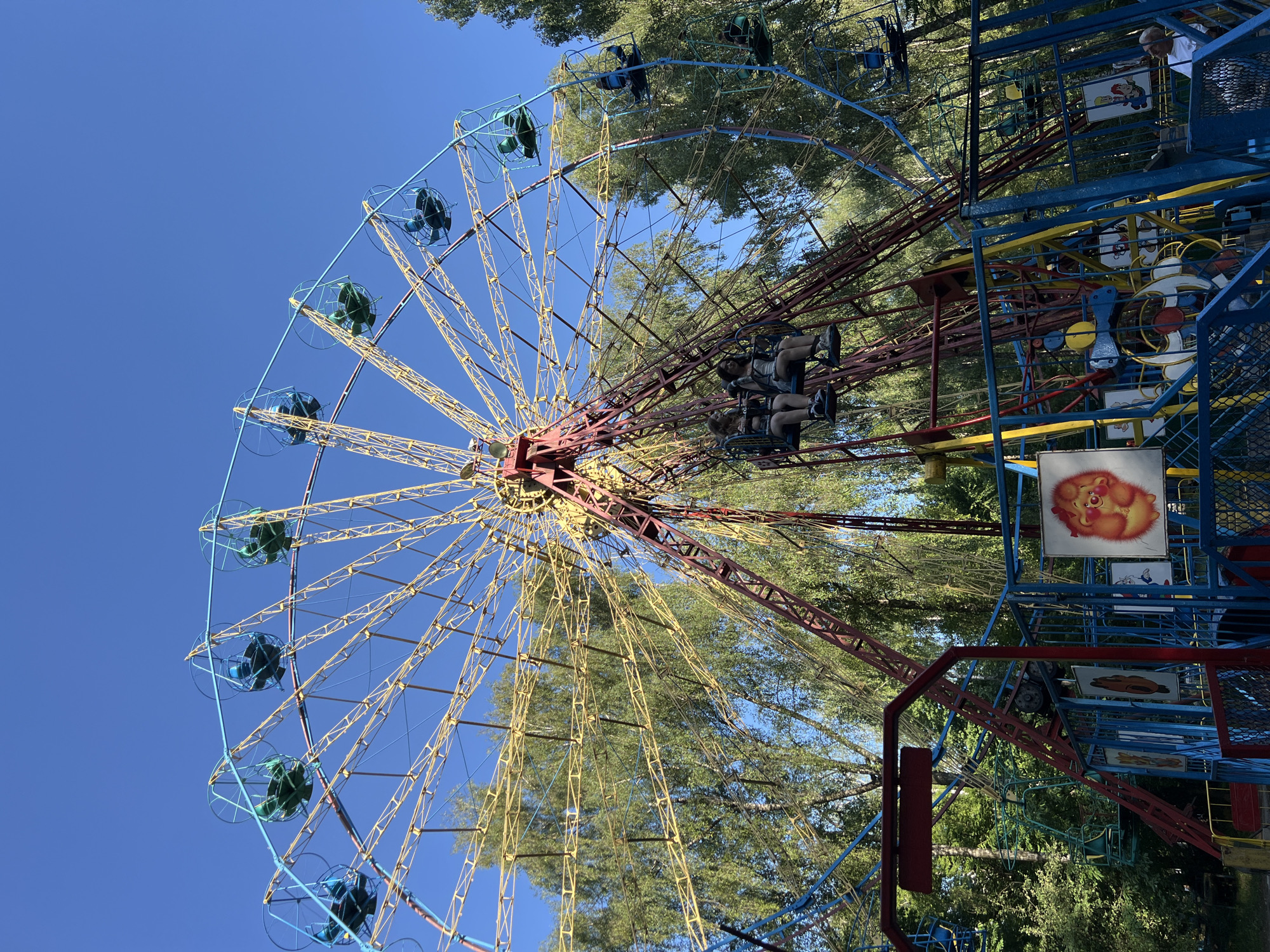 This image depicts a Ferris wheel at an amusement park, likely located in Ternopil, Ukraine, given the provided context. The Ferris wheel is constructed with a predominantly yellow and red metal frame, with blue accents on the passenger cabins. The cabins are enclosed with a blue wire cage and have green seats. Several cabins are visible, some with passengers. In the foreground, there are fenced-off areas of the amusement park, with brightly colored fences and cartoonish signage. The environment is lush with green trees surrounding the Ferris wheel, indicating an outdoor setting. The sky is a clear blue. The image captures a sunny day, likely during daytime hours.