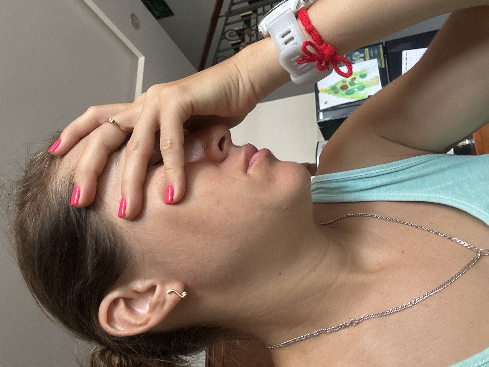 A woman with pink fingernails is seen in a close-up, angled shot. She is wearing a light blue tank top and a silver chain necklace. On her left wrist is a white sports watch with a red string tied around it. She has a nose piercing, and a gold, wave-shaped earring is visible in her left ear. She is touching her forehead with her hand, and the background appears to be indoors. A monitor displaying what looks like a Christmas tree can be seen in the background. It is not possible to confirm the location is in Bac Nha Trang Ward, Vietnam, from the image alone.