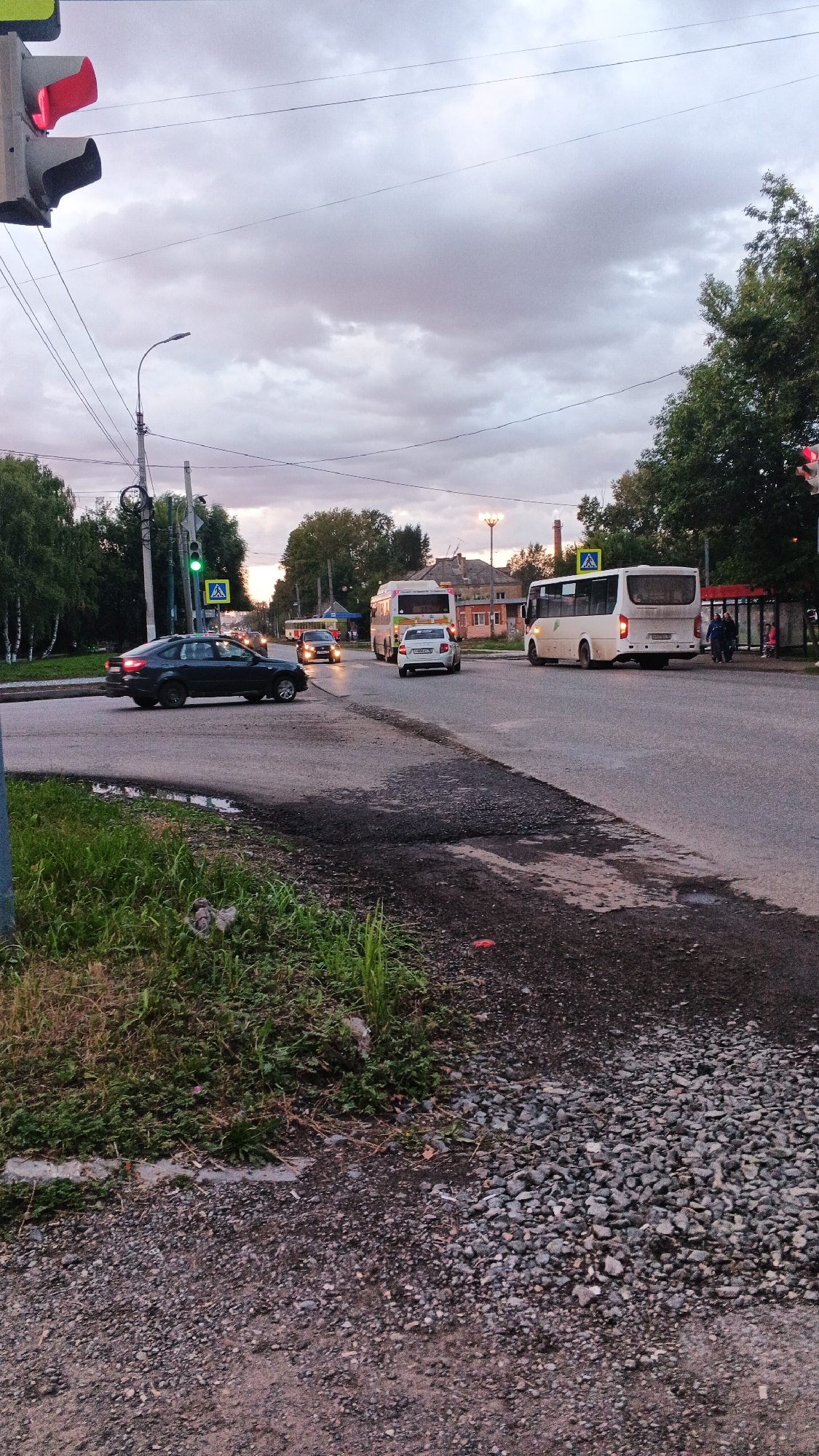 The image captures a street scene in Izhevsk, Russia, likely during the evening with overcast skies. In the foreground, a dark-colored sedan is turning left at an intersection. The traffic light on the left is red. Several buses are also visible on the road, with one white bus in the middle ground and another yellow and white bus further back. Pedestrians are present near a bus stop on the right side of the road. The environment includes trees lining the street, power lines overhead, and buildings in the background, suggesting an urban or suburban setting. The road surface shows signs of wear and patches of gravel.
