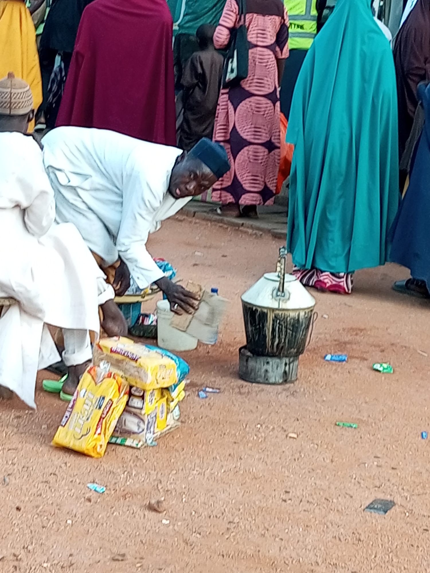 The image depicts a street scene in Gombe, Nigeria, with several individuals present. In the foreground, a man wearing a white garment and a dark cap is bent over, appearing to interact with items on the ground. He is positioned next to a stack of yellow-packaged goods, possibly food items, and a large, dark metal pot sitting on a stove. To his left, another person in a white garment is seated.
In the background, several other people are visible, some in colorful traditional attire. One person is wearing a bright pink and purple patterned dress, and another is covered in a large teal-colored garment. The ground appears to be dusty or sandy. The overall impression is of a bustling outdoor environment, potentially a market or a gathering place.