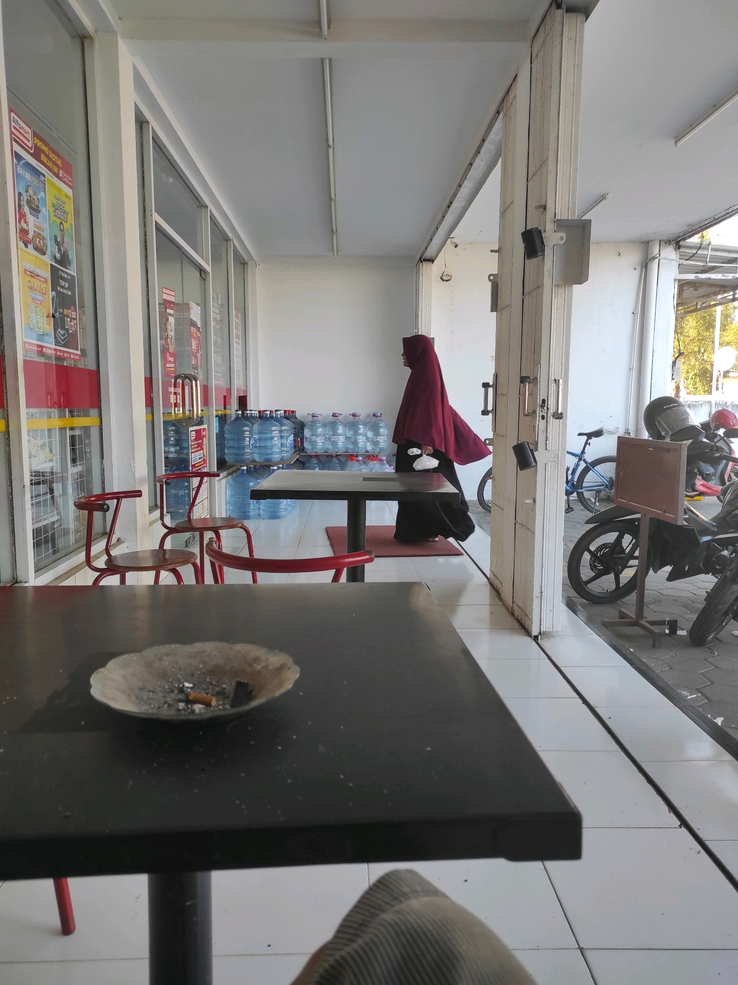 The image depicts an outdoor seating area in front of a store, likely in Purwodadi, Indonesia. In the foreground, a dark rectangular table with an ashtray containing cigarette butts is visible. In the mid-ground, red chairs and a table are arranged on white tiled flooring. Behind this, a person wearing a maroon hijab and dark clothing walks away from the camera. The store's interior, featuring rows of bottled water, is visible through large glass windows. Further back, a blue bicycle and motorcycles are parked. The environment appears to be a public space with the store likely a minimarket or convenience store.