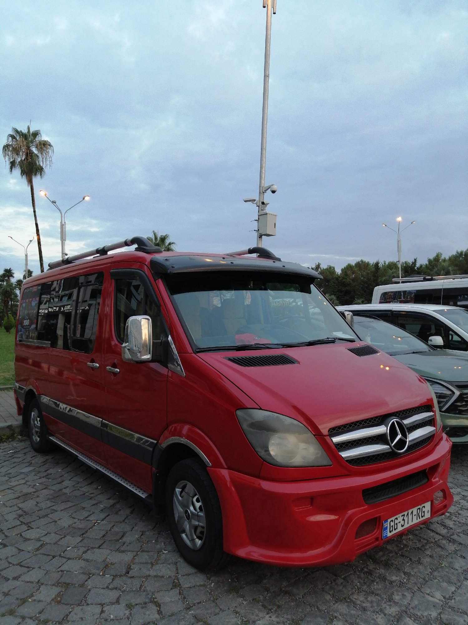 A red Mercedes-Benz Sprinter van is parked on a cobblestone surface. The van has a silver roof rack and chrome trim. The front of the van features a Mercedes-Benz logo, a silver grille, and a red bumper with a license plate that reads "GG-311-RG". To the right of the van, a light-colored SUV is partially visible. In the background, a white bus with "ROAD" written on its side is parked. Palm trees and other vegetation line the background, suggesting a location with a temperate climate. Several streetlights are present, and a security camera is mounted on a pole. The sky is overcast.