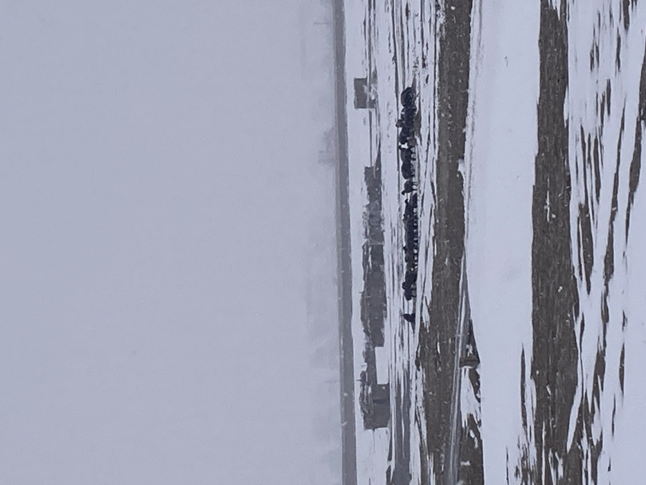 The image displays a vast, open landscape in Khanai Baba, Pakistan, under an overcast grey sky. The ground is partially covered with irregular patches of snow, interspersed with exposed dark brown earth. In the mid-ground, a herd of approximately 20-25 dark-colored animals, consistent with sheep or goats, is grouped together. To the left of this herd, a single individual, dressed in dark clothing, is seated or crouched directly on the snow-dusted ground. Behind the animals, several low-profile, rectangular structures, appearing to be constructed from earthy materials, are visible. A long, dark, linear feature stretches horizontally across the distant mid-ground, possibly an embankment or distant wall. Faint, dark outlines of hills or mountains are discernible on the horizon, partially obscured by atmospheric haze or snowfall.