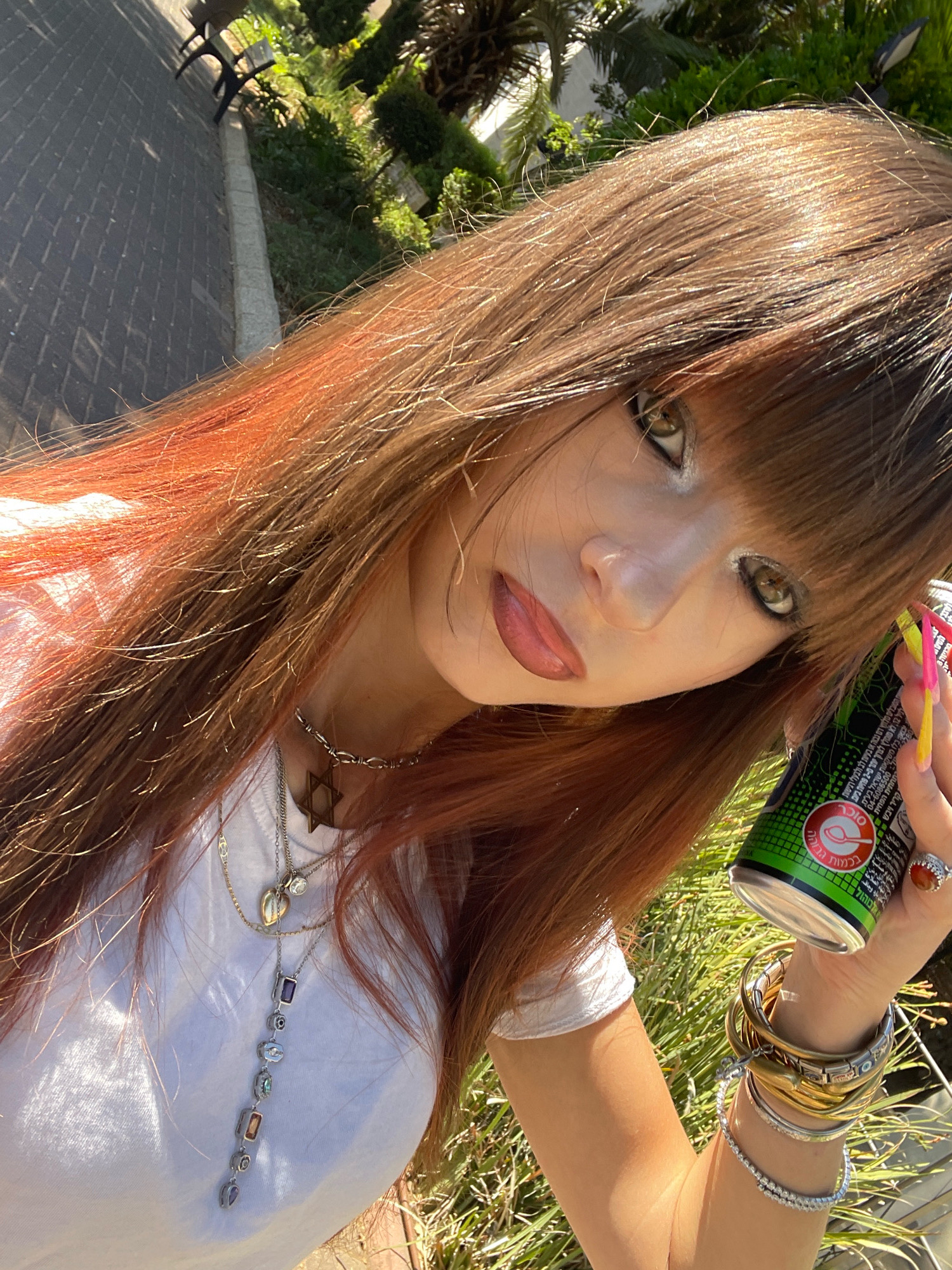 The image shows a person's upper body and face, with their head tilted to the right. They have long, auburn hair with some reddish-orange highlights, and bangs that partially obscure their eyes. The person is wearing a white t-shirt and multiple necklaces, including a prominent Star of David pendant and a long necklace with colorful stones. Their eyes appear to be green and they are wearing noticeable eye makeup. On their right hand, they are holding a green can and wearing a ring with an orange stone. Their nails are brightly colored, with yellow and pink tips. The person is surrounded by greenery and some paved ground with benches in the background. The lighting suggests it is daytime. Given the text on the can, which appears to be in Hebrew, and the presence of palm trees and other vegetation, the location is likely Raanana, Israel.