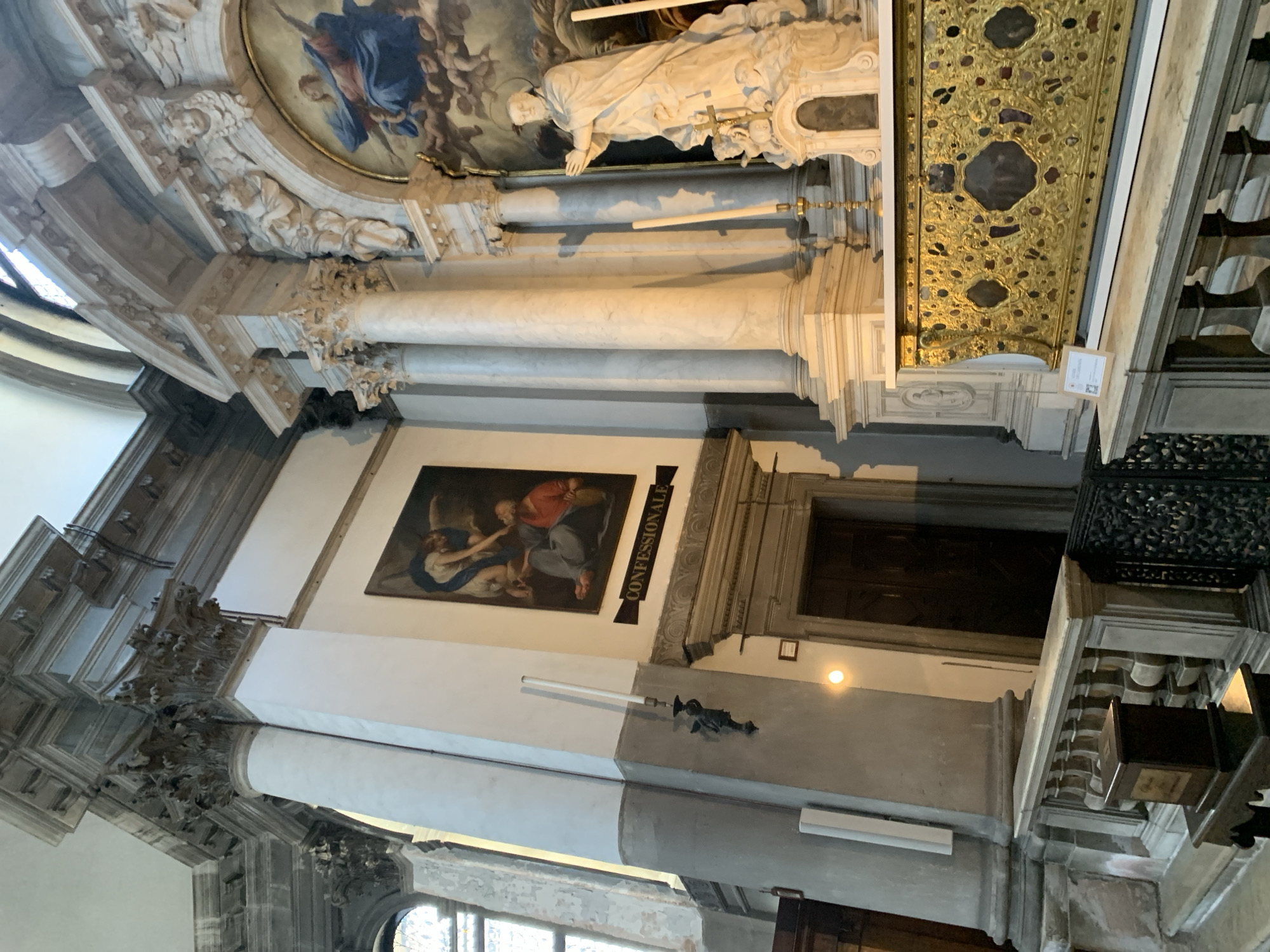 The image shows an interior view of a church in Venice, Italy, likely a sanctuary or chapel. The scene is dominated by ornate architectural elements, including large marble columns with Corinthian capitals, a decorative altar covered in gold leaf and intricate patterns, and a large painting of religious figures. To the left, a dark wooden door is visible, possibly a confessionale, above which hangs a painting depicting an angel assisting a kneeling man. A statue of a robed male figure with clasped hands stands to the right of the altar, in front of a framed religious painting. The space is dimly lit, with light streaming in from a window on the left and from some interior lighting fixtures. The overall impression is one of grandeur and solemnity.