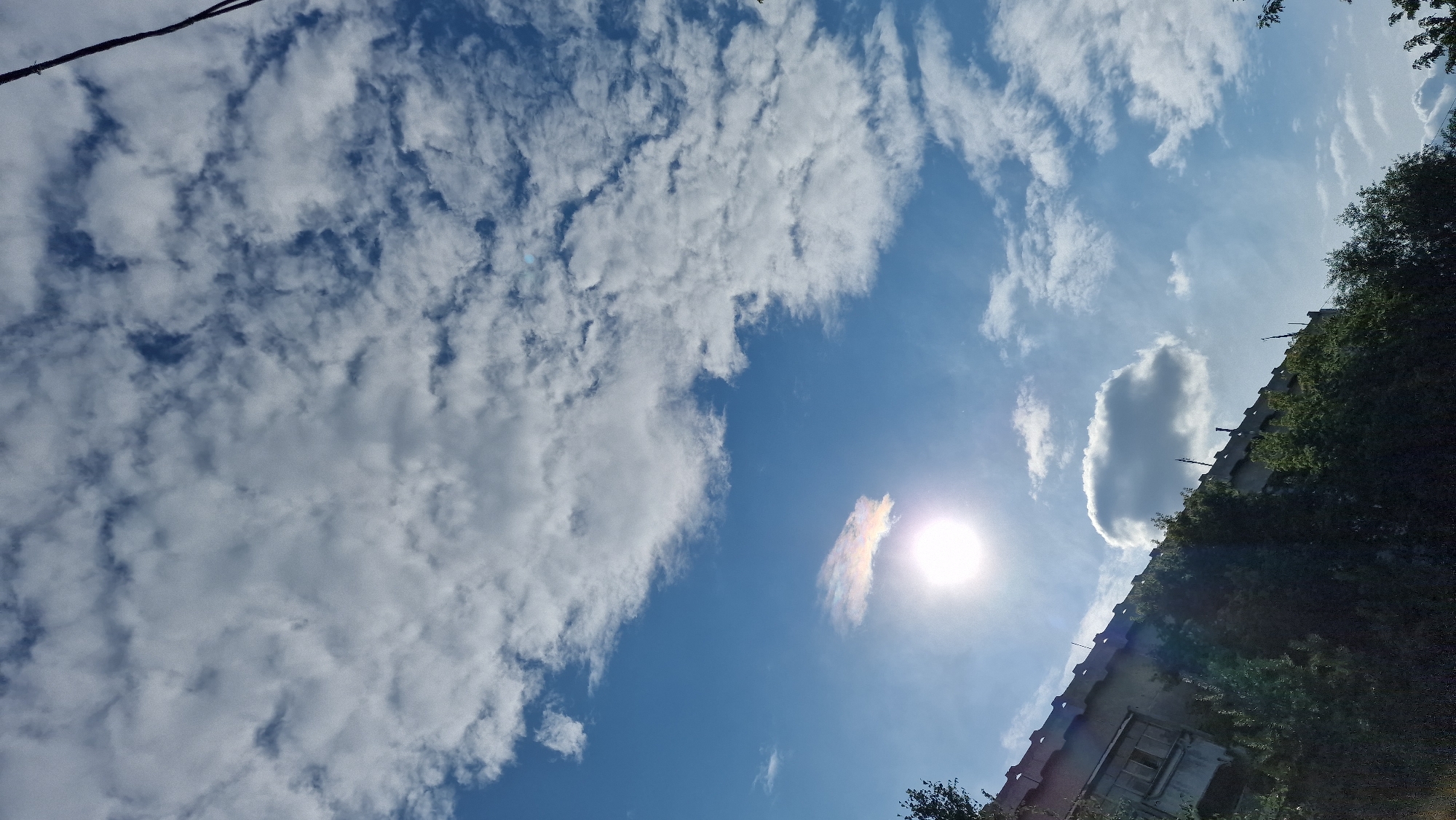 The image shows a cloudy sky with the sun partially visible, casting a bright light. There is a subtle rainbow-like halo around the sun, suggesting a sun dog phenomenon. In the foreground, the upper part of a building with a textured, concrete facade is visible, with a row of green trees and bushes below it. A telephone wire runs diagonally across the top right corner of the frame. The scene appears to be an exterior view of an urban area.