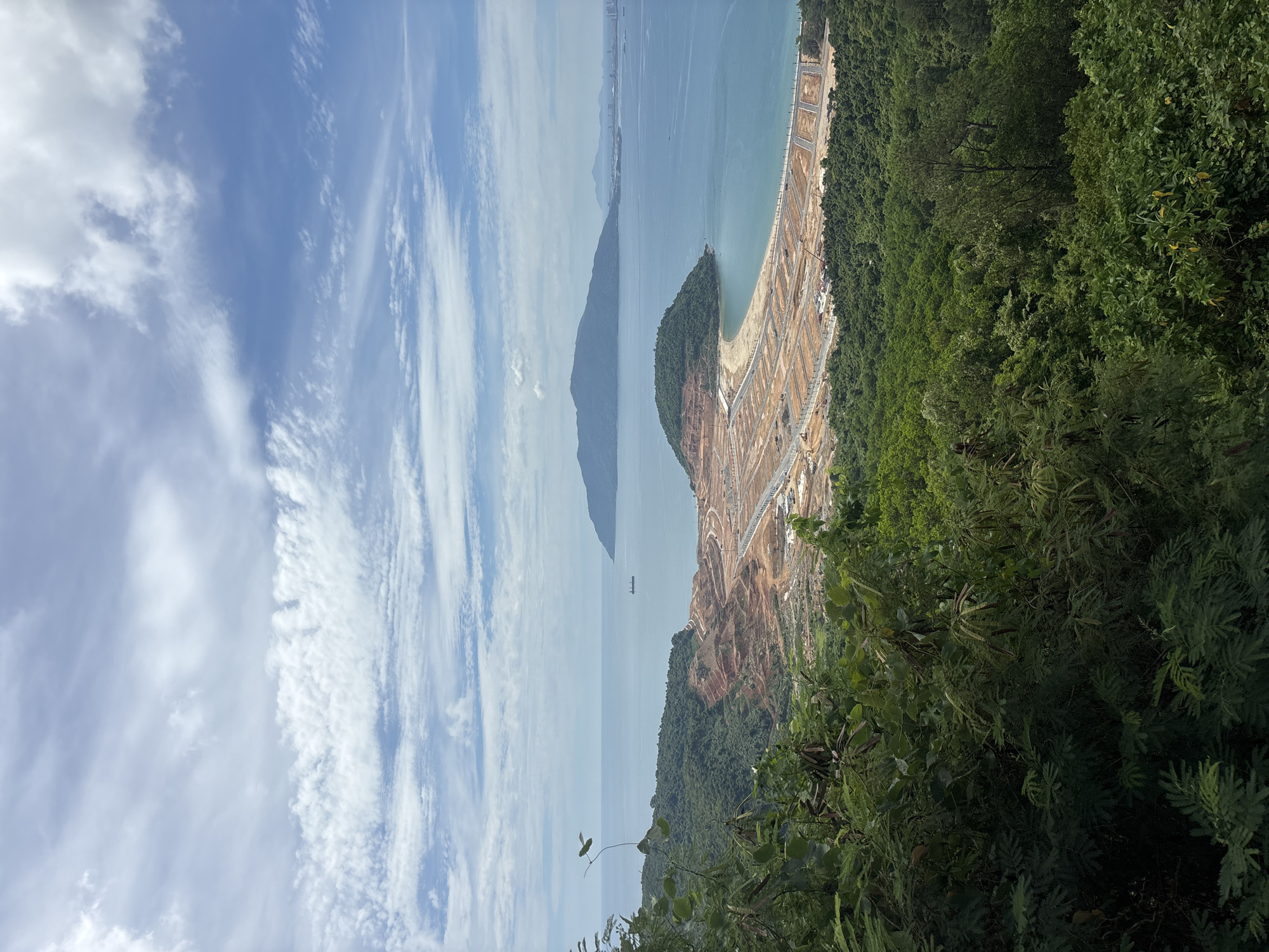 The image captures a panoramic view from a verdant hillside overlooking a developing coastal area in Đà Nẵng, Vietnam. In the foreground, lush green foliage dominates, partially obscuring the view. Below, a significant construction site is visible on a curved bay, characterized by cleared land, graded earth in various shades of brown and orange, and a grid of newly laid roads. Construction equipment, including cranes, can be spotted within the site.
The bay itself is a striking turquoise, contrasting with the construction zone. Further out in the sea, a lone cargo ship is visible in the distance. Across the water, a large, mountainous island rises, with a smaller, rounded island or promontory situated closer to the construction area. The horizon is dotted with distant coastal features and possibly part of the Đà Nẵng cityscape. The sky above is a dynamic mix of blue and white clouds, with wispy cirrus clouds stretching across a significant portion of the frame. The overall scene depicts a transition from natural landscape to urban development.
