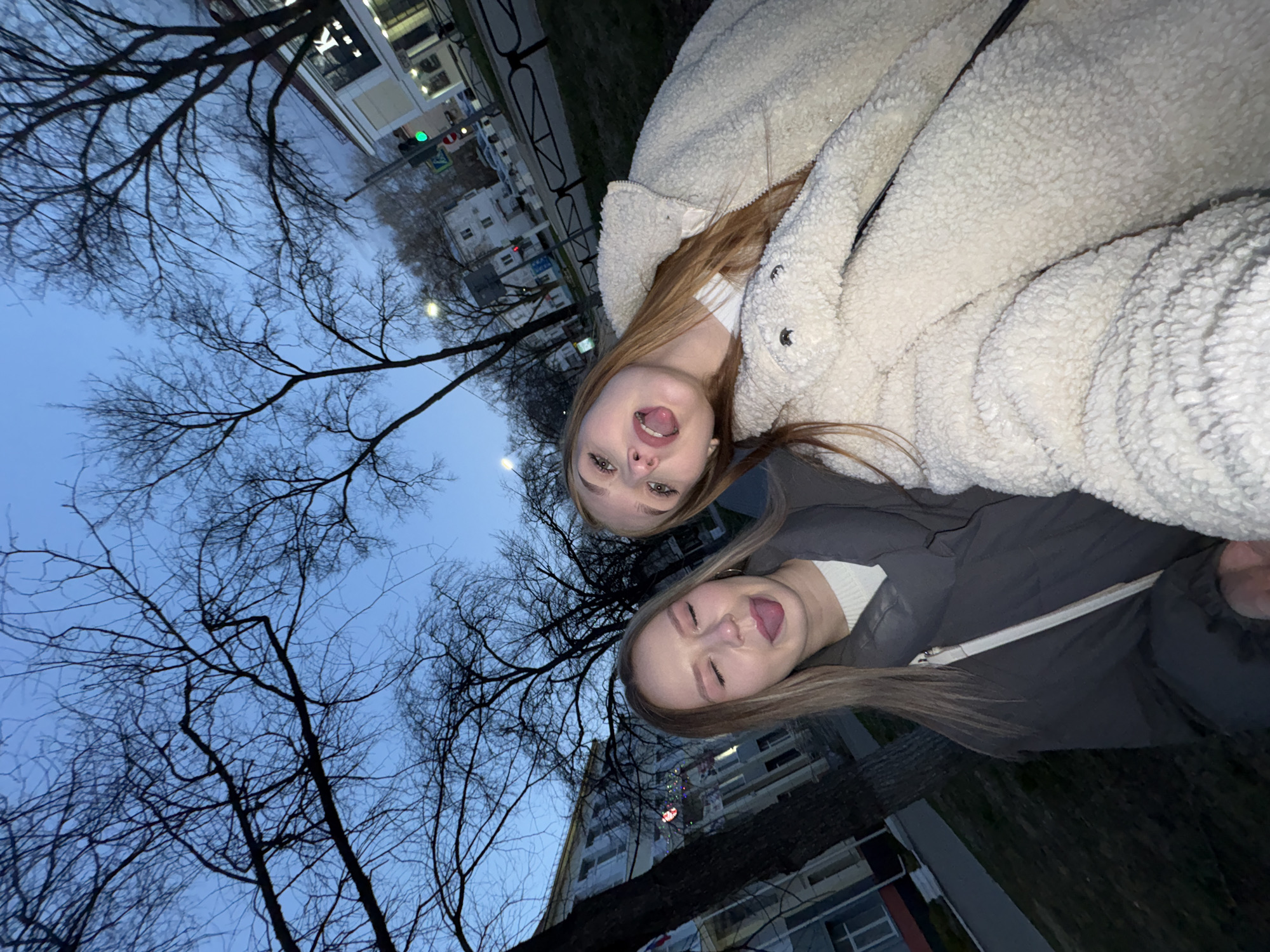 Two young women are captured in a playful, low-angle selfie outdoors, likely during dusk or early evening. The sky, a deep blue, fills the upper portion of the image, silhouetting numerous bare tree branches, suggesting a colder season like late autumn, winter, or early spring.
The woman in the foreground, positioned on the right side of the frame, has long, light brown hair and is wearing a thick, textured, cream-colored teddy coat over a white top. She has a wide-open mouth expression, showing her tongue and teeth, indicative of laughter or a playful shout. Behind her and slightly to the left, the second woman, also with long, light brown hair, is wearing a dark grey or black puffer jacket over a white ribbed top. She is sticking out her tongue with a playful, squinted-eye expression.
In the background, urban elements are visible. On the far left, a multi-story building features a distinct white letter "K" on a dark sign. A green traffic light and a pedestrian crossing sign are also discernible near a street or road. Other buildings and what appears to be a fence or railing line the area, with patches of grass or ground visible below the trees. The overall mood is lighthearted and joyful. The location context for this scene is Chaikovsky, Russia.