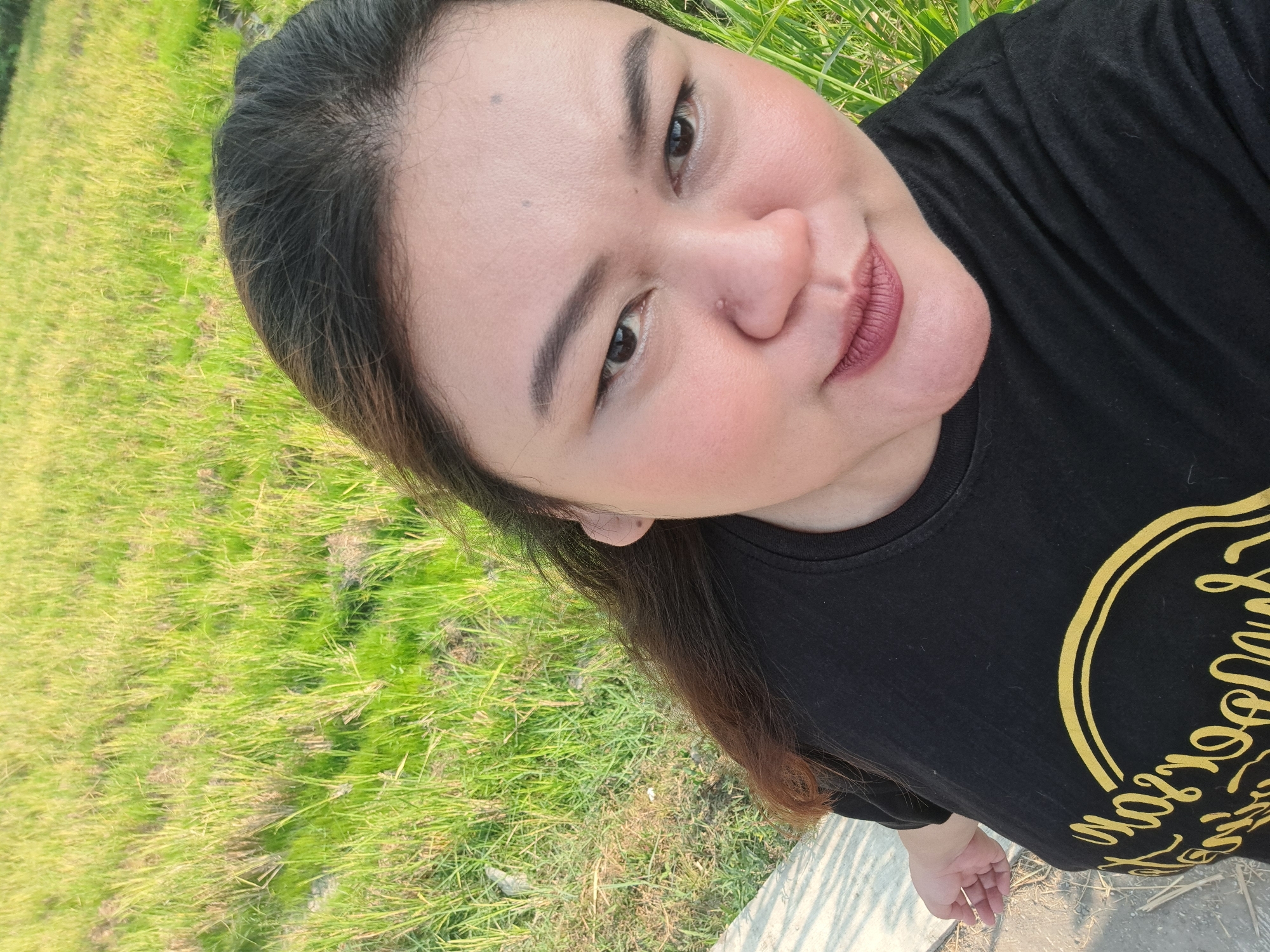 A selfie is taken from a high angle, showing a woman with dark hair and makeup, wearing a black t-shirt with yellow lettering. She is positioned in the foreground, with a green grassy field stretching out behind her. A concrete path is visible to her left. The lighting suggests it is late afternoon. The image captures a portrait of the woman against a natural backdrop.