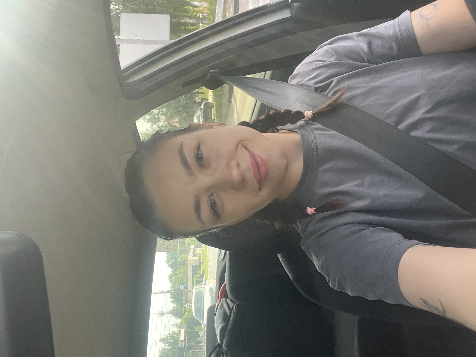 A young woman with braided hair and wearing a grey t-shirt is sitting in the driver's seat of a car. She is smiling and looking at the camera. The interior of the car is visible, including the dashboard and the seatbelt. Outside the car, a street scene with trees, buildings, and other cars can be seen. The image appears to have been taken during the daytime on a clear day. There is a faint glow of sunlight, possibly indicating a late afternoon or early morning setting. The woman has a tattoo on her left arm. The overall impression is a casual selfie taken while in a vehicle, likely in a suburban or semi-urban environment. It is not possible to definitively determine the specific location within Chaikovsky, Russia from the image alone.
