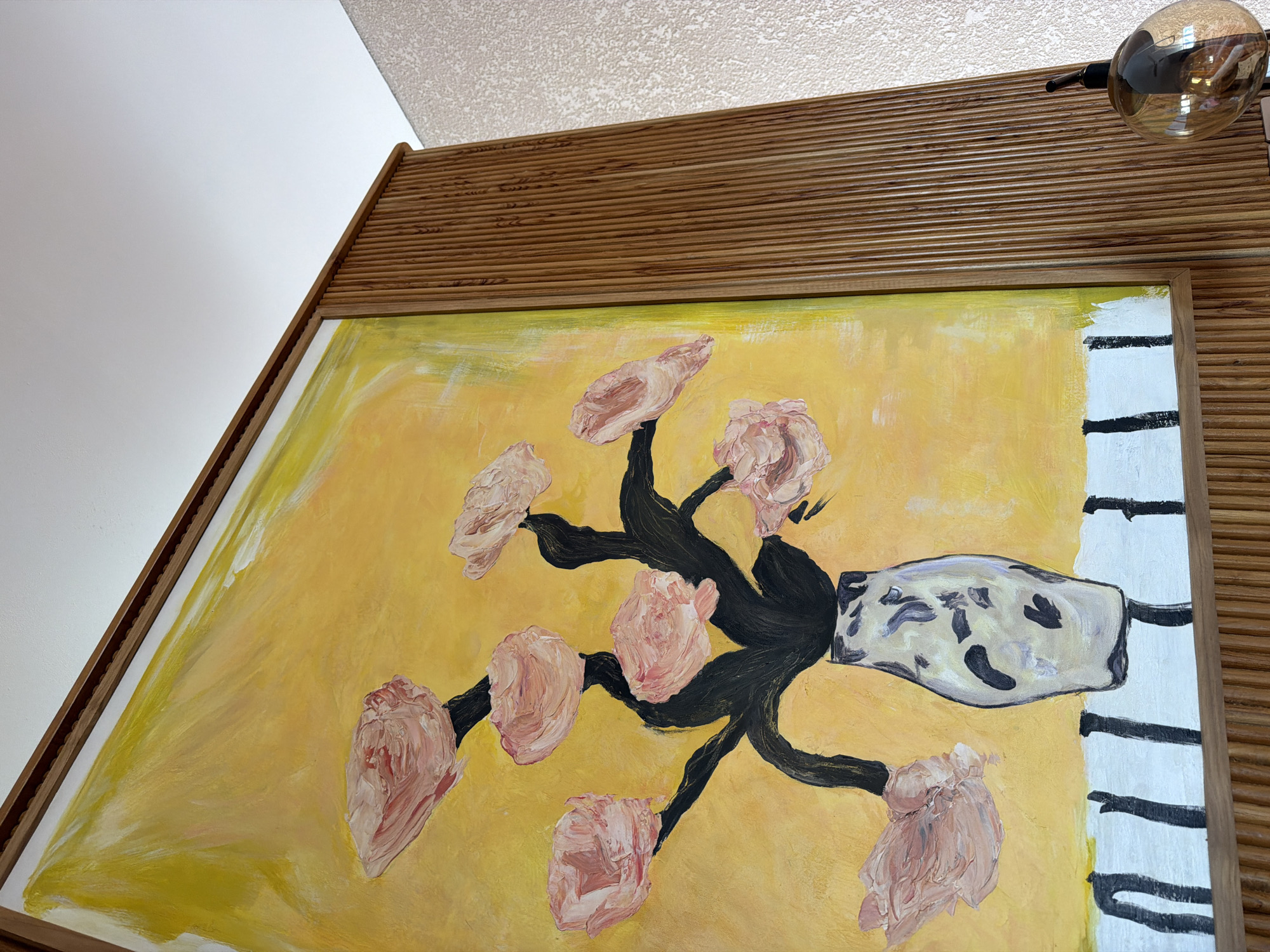 A close-up shot focuses on a framed painting with a yellow background. The painting depicts stylized flowers with prominent, thick brushstrokes in shades of pink and peach, connected by dark, winding branches. A vase with a mottled pattern in shades of gray and black rests on a white surface with black horizontal stripes. The frame is composed of light brown wood, with a fluted or ridged texture. The top portion of the frame is visible, extending to the ceiling. A partially visible light fixture with a transparent globe is positioned above and to the right of the frame. The location of the shot appears to be in Mengwi, Indonesia.