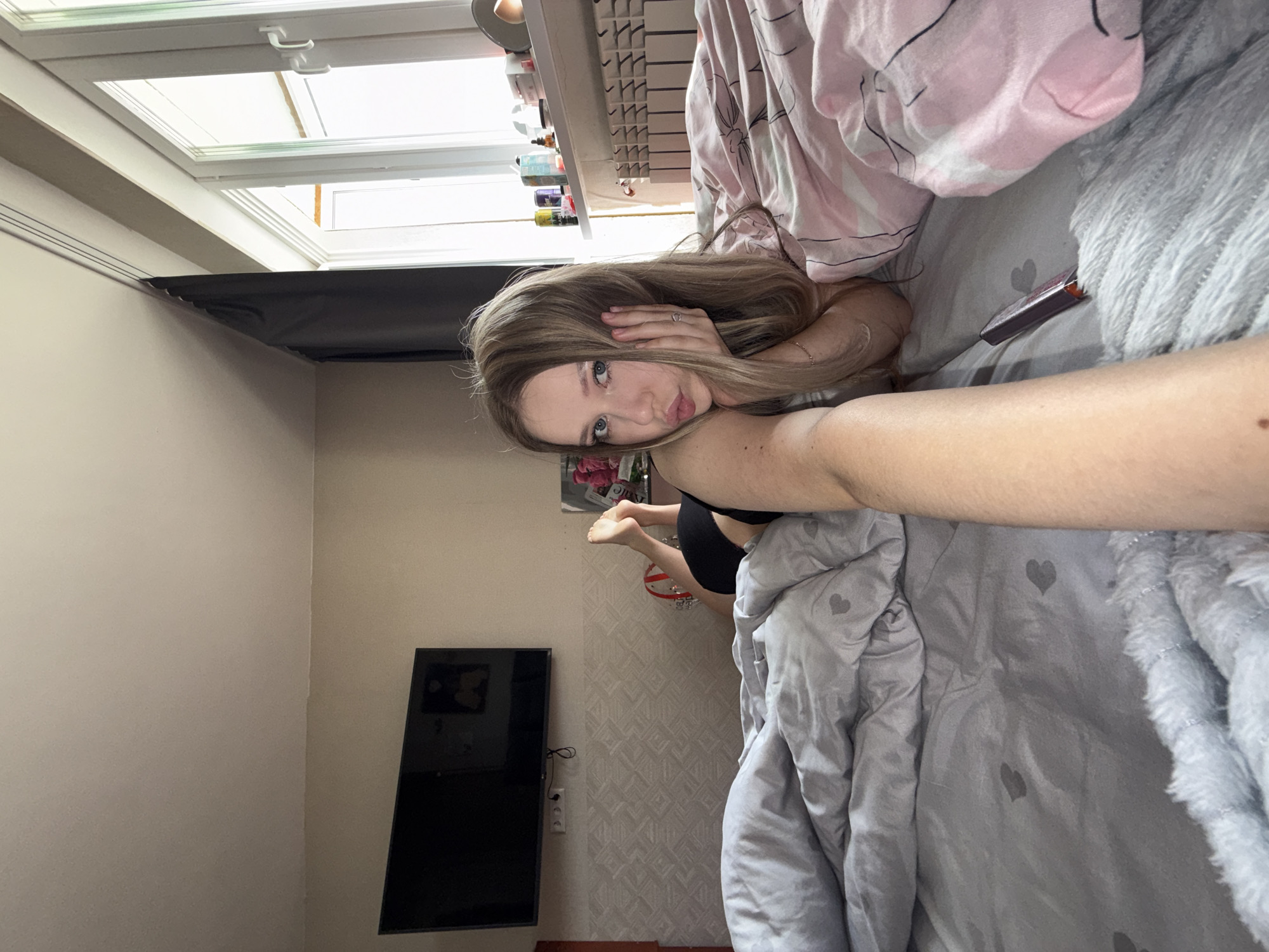 A young Caucasian woman with long, blonde hair and blue eyes is lying on a bed, facing the camera. She is wearing black underwear. Her left arm is extended towards the camera, and her right hand is propped under her chin, with her fingers resting on her cheek. She is making a pouty face. The bed is covered with grey and pink bedding. In the background, there is a television mounted on the wall, a window with blinds, and a radiator. The room appears to be a bedroom. The image is taken from a low angle, with the woman's arm dominating the foreground. There is a general sense of casualness and intimacy in the scene.