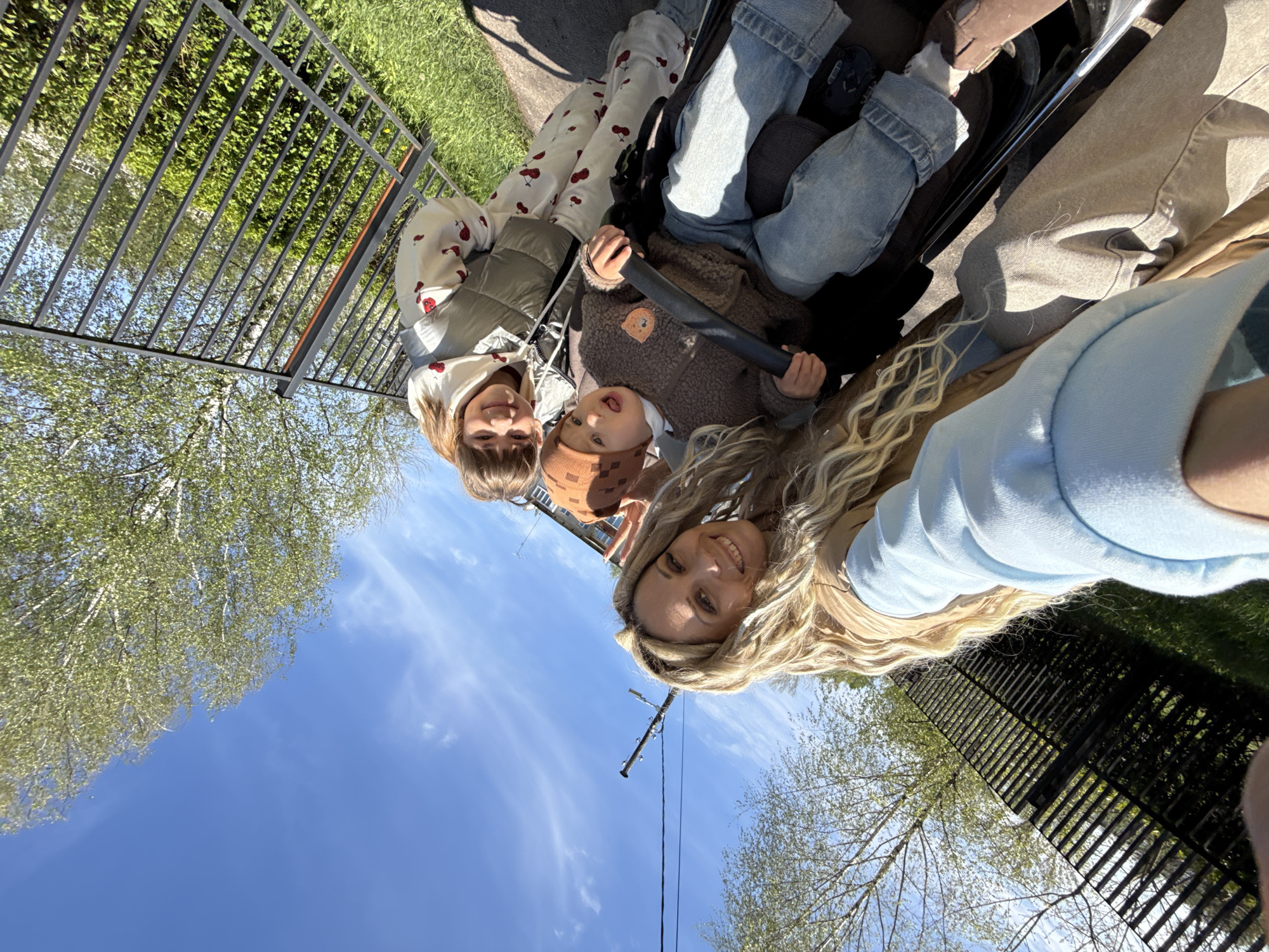 The image depicts a selfie taken outdoors with a woman and two children. They are positioned in what appears to be a stroller. A black metal fence is visible behind them, along with trees and a blue sky. The specific location is Rusanovo, Russia. There is also a utility pole visible in the image. The people are smiling and interacting, indicating a pleasant scene.