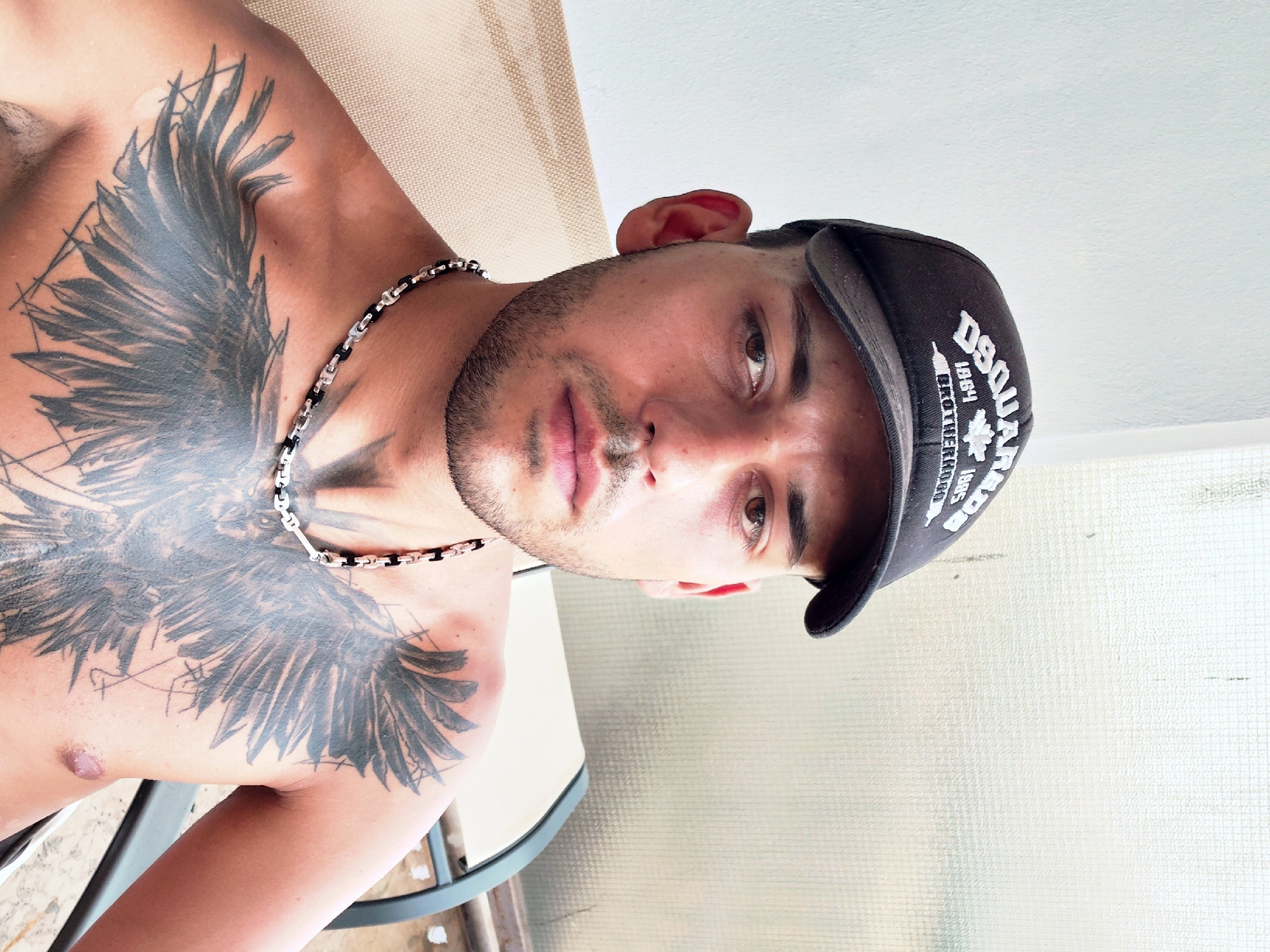 A man wearing a black baseball cap and a silver and black chain necklace is photographed from the chest up, sitting outdoors in what appears to be a patio or balcony setting. The man has a prominent black tattoo of bird wings covering his chest and upper torso. He has short, dark hair and a light beard. The background consists of a white wall and a textured, translucent screen or window. The lighting suggests it is daytime. There is no indication of the specific location within the Municipal Unit of Aegio, Greece.