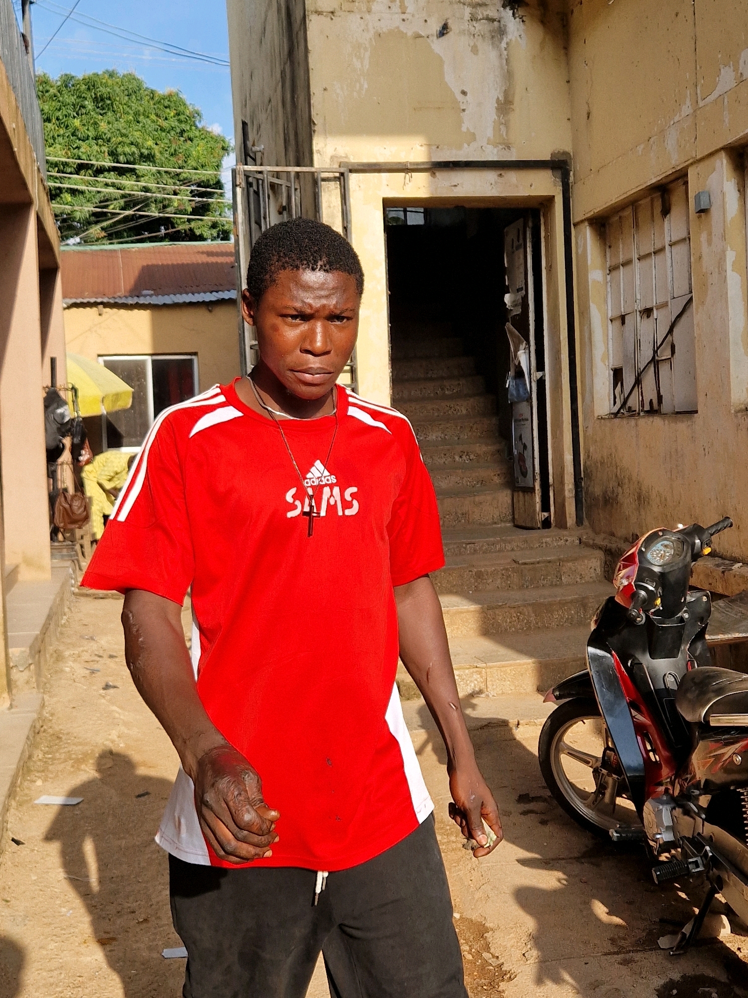 A young Black male stands in the foreground of an urban alleyway. He is wearing a red t-shirt with white stripes on the sleeves and black track pants. A silver necklace with a cross pendant is visible around his neck. His left arm appears injured, with visible abrasions and a wound on his hand. He is holding a small, light-colored object in his right hand. Behind him, a building with peeling yellow paint and a dark doorway with stairs leading up is visible. A red and black motorcycle is parked to his right. The ground is dirt and concrete, with shadows cast by the subjects and overhead wires. In the background, trees and a blue sky are visible.