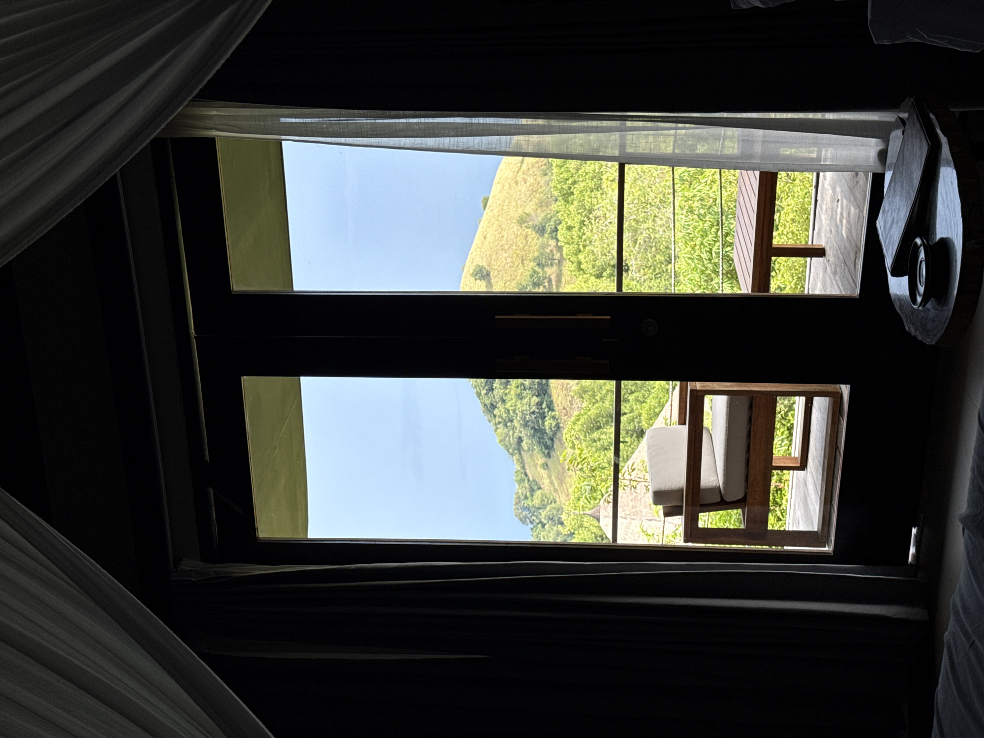 View from within a dimly lit interior, possibly a room, through an open door or window. The scene outside features a grassy hillside with green vegetation under a clear sky. A wooden deck or balcony is visible just beyond the opening, containing a chair with light-colored cushions. A round table with objects on it is located near the right side of the opening. The location is potentially in Unknown, Indonesia.