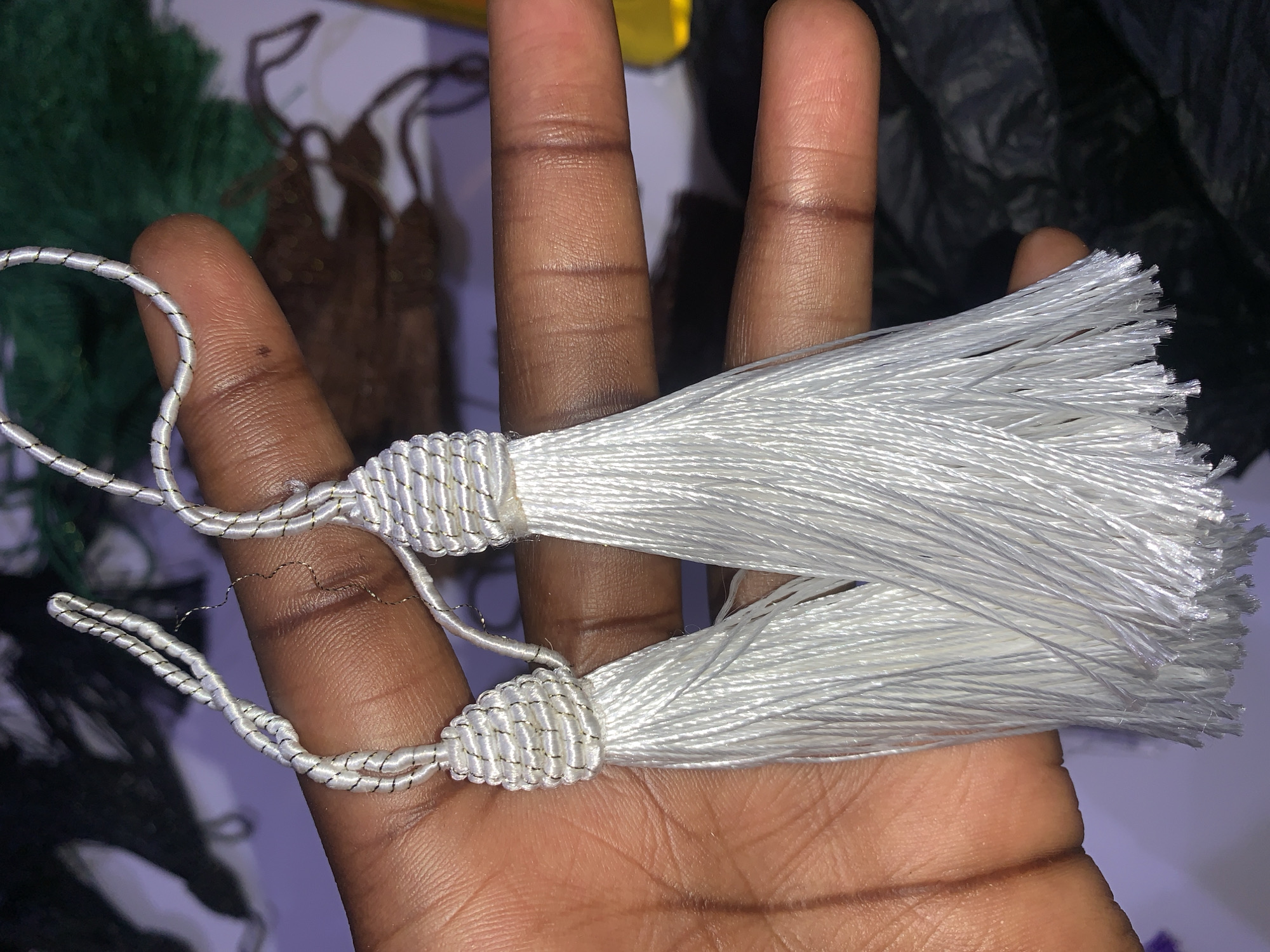 In Gura Top, Nigeria, a close-up shot shows a hand holding two white tassels. The tassels are made of thin, white threads and have a decorative, braided top. The hand, with dark skin and visible fingerprints, is positioned behind the tassels, supporting them. The background is blurred and dark, with hints of green and brown objects, suggesting an indoor or possibly outdoor setting with some decorative elements. The lighting is artificial, casting shadows and highlighting the textures of the tassels and skin.
