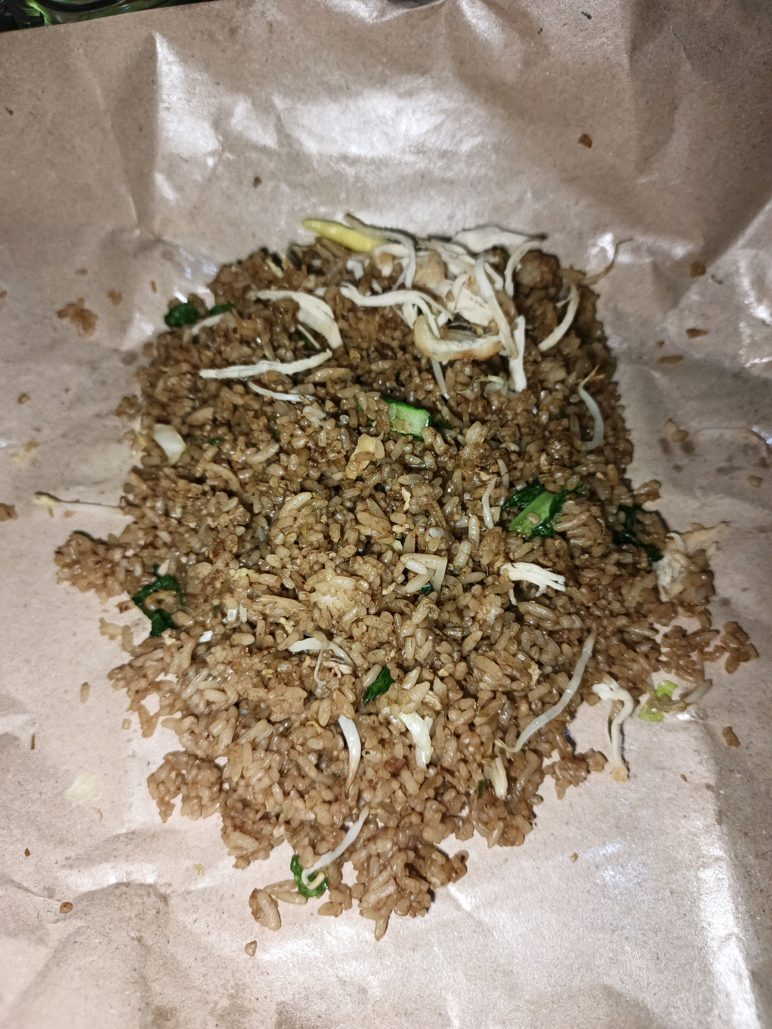 In Ketegan, Indonesia, a pile of nasi goreng, or Indonesian fried rice, is presented on a brown paper wrapper. The rice is a rich brown color, indicating it has been stir-fried with soy sauce and other seasonings. Scattered throughout the dish are shredded chicken, bean sprouts, and chopped green onions, adding texture and visual appeal. The paper wrapper is slightly crumpled, suggesting the food may have been recently served or is intended for takeaway. The lighting in the image is warm and somewhat dim, creating a cozy and inviting atmosphere.