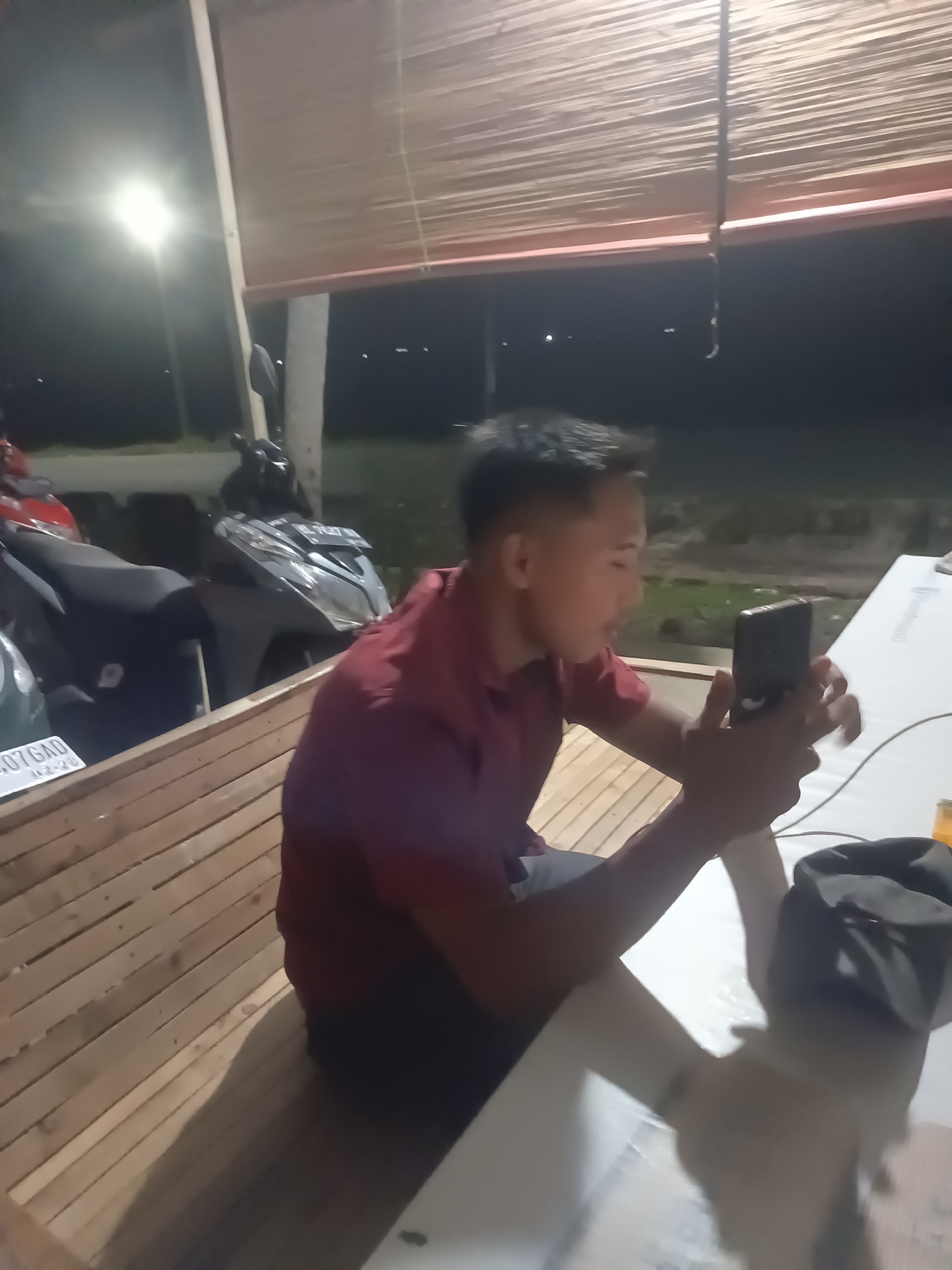 A person is sitting on a wooden bench and looking at their phone. The person is wearing a red shirt and dark pants. They are in an outdoor seating area with a bamboo roof. There are several motorcycles parked in the background. The image appears to have been taken at night, with a bright light source to the left of the frame. The location is in Lhokseumawe, Indonesia.