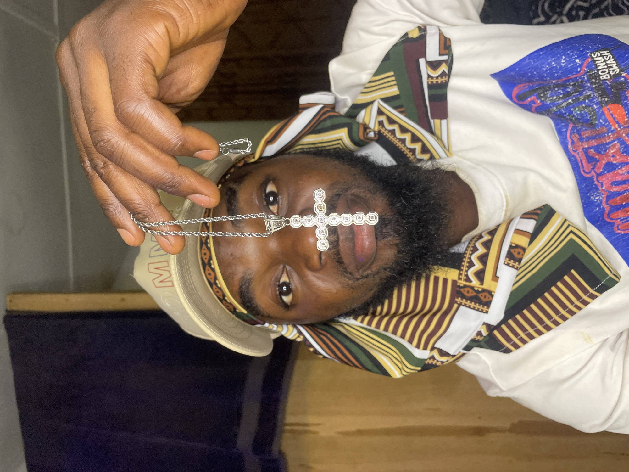 A man is depicted in a close-up shot. He has dark skin, a full beard, and wears a head covering with geometric patterns. He is also wearing a light-colored baseball cap. A silver chain necklace with a large cross pendant is held up near his face by his hand. The location appears to be indoors. The city of Jos, Nigeria is not evident in the image.