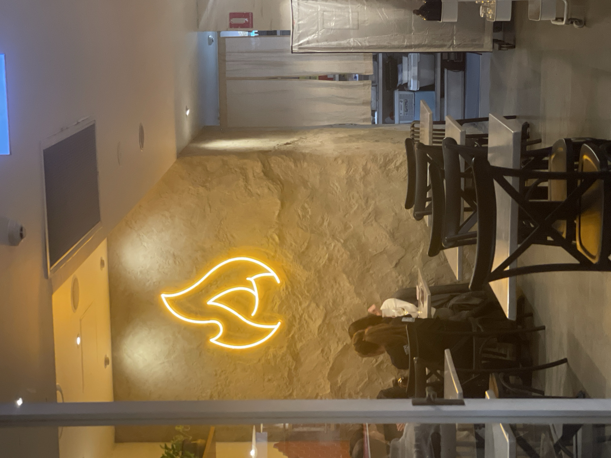 The image shows the interior of a restaurant. A neon sign shaped like a flame is visible on a textured, light brown wall. Several tables and chairs are arranged in the dining area, some occupied by diners. In the background, white curtains hang over a doorway, and some storage shelves are visible. The lighting is warm, primarily from overhead lights and the neon sign. The setting appears to be a casual dining establishment. There is no indication of the city of Brisbane, Australia, within the image itself.