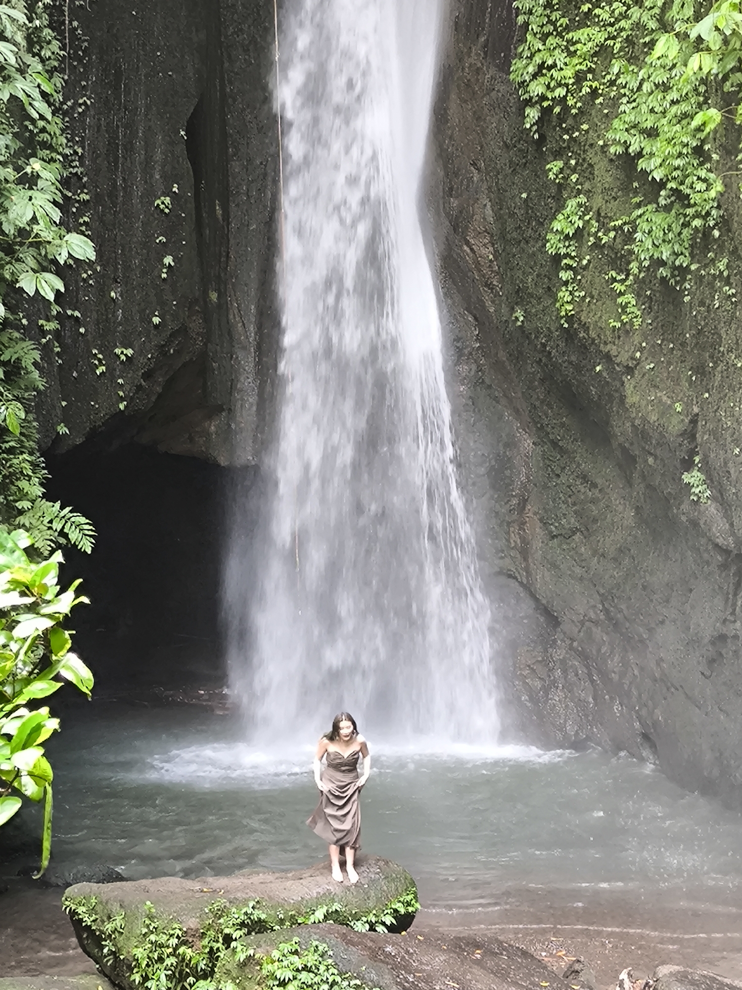 A woman stands in front of a waterfall at Mekarsari, Indonesia. She is in the center, facing forward, wearing a strapless brown dress and white shoes. The waterfall cascades down a rock face into a pool of water where the woman is standing. The surrounding area is covered in green foliage. The waterfall is framed by rocky cliffs covered in vegetation. There is a cave behind the waterfall.