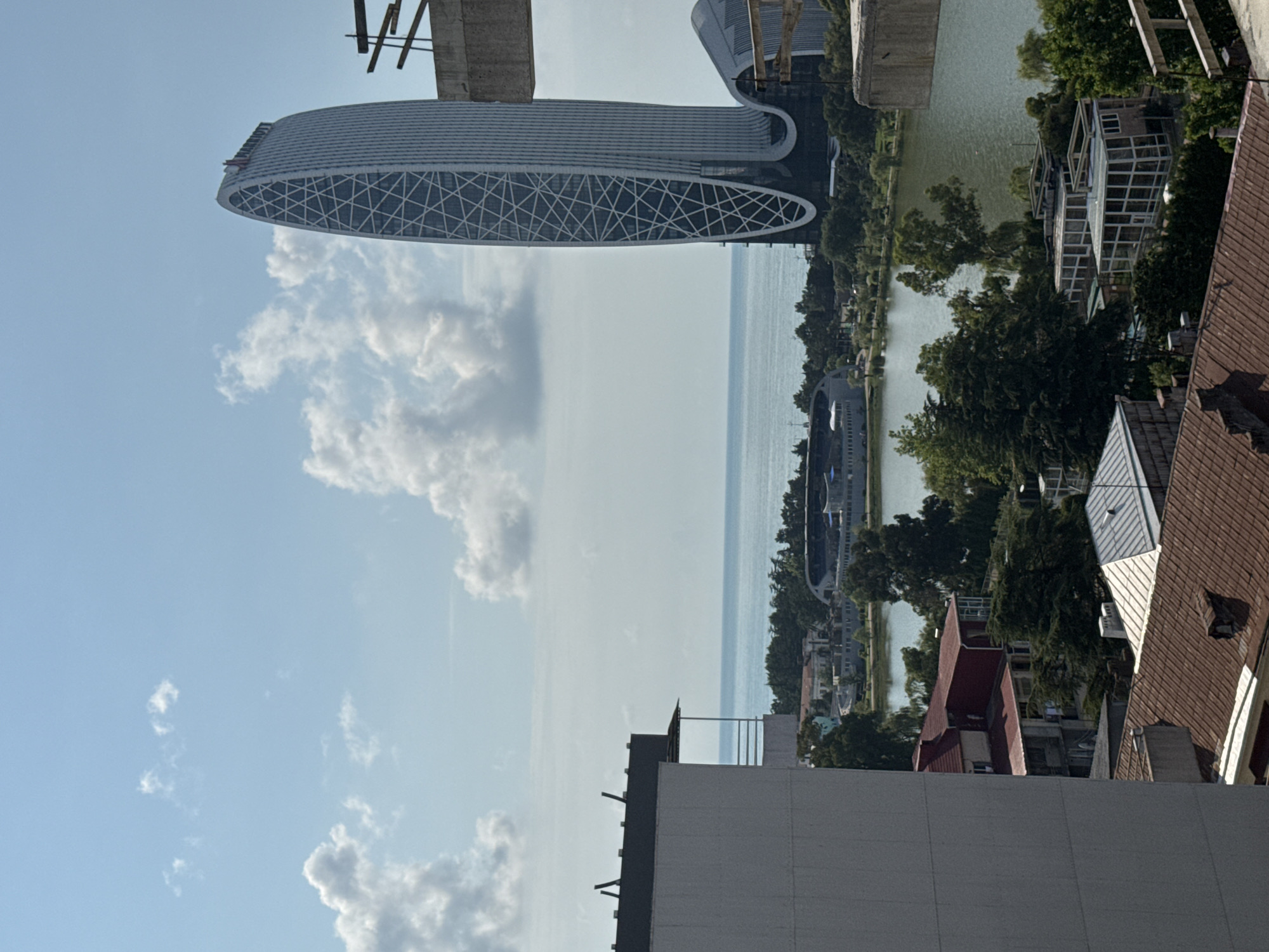 The image shows a panoramic view of a city skyline by the sea. In the foreground, a tall, modern building with a distinctive oval shape and a grid-like facade dominates the right side of the frame. The building has the "Marriott" logo at its apex. To the left of this building, part of another construction site with scaffolding and concrete structures is visible.
Below the modern building, a body of water, likely a lake or bay, is present, with a stadium or large complex visible across it. A treeline separates the water from the buildings. In the mid-ground, a cluster of residential buildings with red roofs are visible. The sea stretches out to the horizon under a partly cloudy sky. The overall scene appears to be a coastal urban environment, possibly during daylight hours.