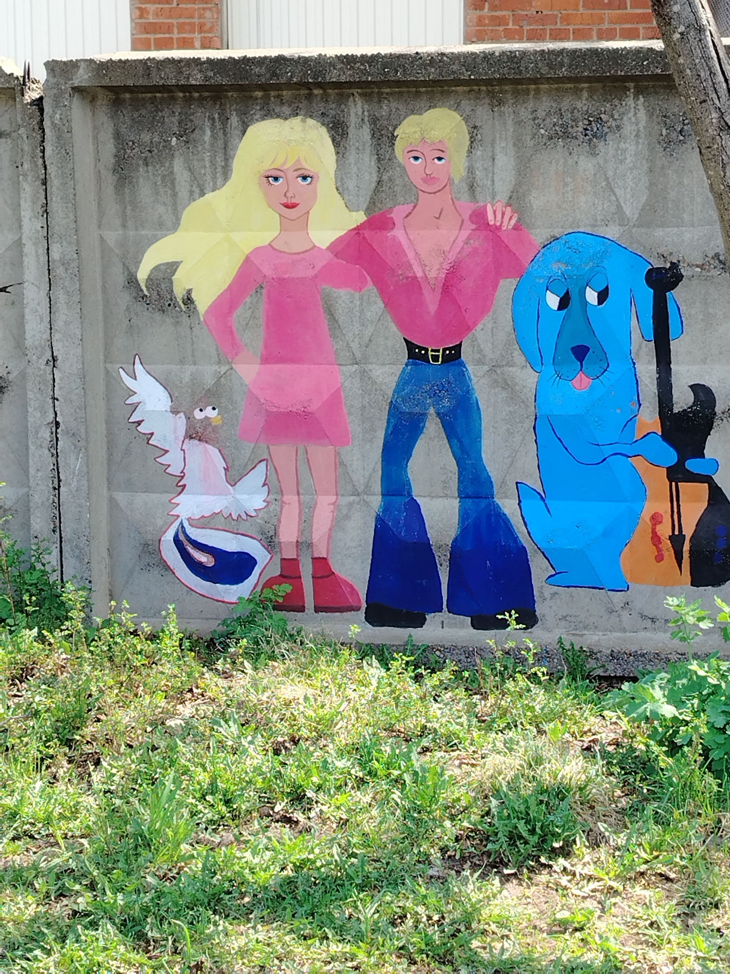 The image depicts a section of a concrete wall, likely a fence, upon which a mural has been painted. The mural features a blonde-haired woman in a pink dress, standing beside a blonde-haired man in a pink shirt and blue jeans. The man has his arm around the woman. To the left of the couple is a painted bird, possibly a dove or pigeon. To the right of the couple is a blue dog, holding a black and brown guitar. The wall is grey and weathered. Below the wall is a patch of green grass and weeds. Based on the location listed, this image appears to be in Noviy, Russia.