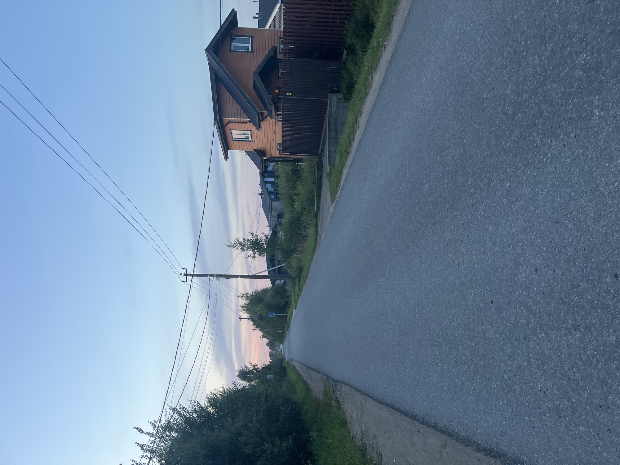 The image depicts a rural road at dusk in the Babigonskaya valley, Russia. The road is paved and runs through a sparsely populated area with trees and vegetation on either side. Several utility poles with wires are visible along the road, indicating power and communication lines. On the right side of the road, a two-story wooden house with a dark roof and light-colored window frames is present, surrounded by a dark wooden fence. Further down the road, more houses and trees can be seen. The sky is a soft gradient of blue and pink, characteristic of twilight. There are no people or vehicles visible in the image.