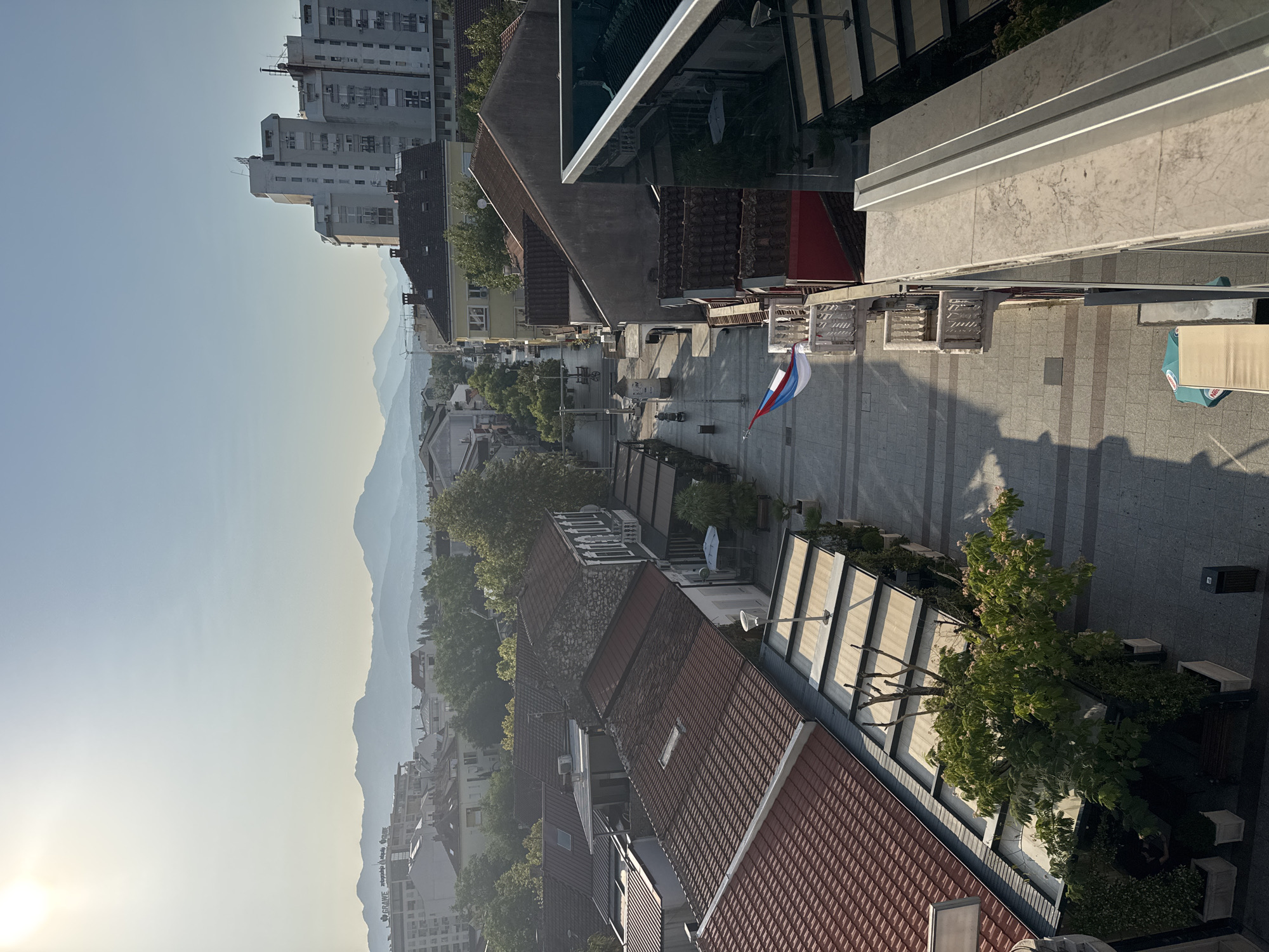 An eye-level, high-angle medium shot captures a street scene in an urban environment during the day. The sun, positioned high in the sky on the left, casts a bright light that creates a hazy atmosphere and long shadows. The street is paved with grey tiles and is lined with buildings on both sides. Some buildings have awnings extending over outdoor seating areas, suggesting cafes or shops. A flag, colored white, blue, and red, is visible on a balcony on the right side of the street. The street extends into the distance, where a row of mountains is silhouetted against the sky. The architecture appears to be a mix of older buildings with tiled roofs and more modern, multi-story structures. Trees and greenery are interspersed among the buildings, adding a touch of nature to the urban landscape. A few people are visible walking in the distance, and a bicycle is parked on the left side of the street. The overall impression is one of a bustling yet serene city street bathed in warm sunlight.