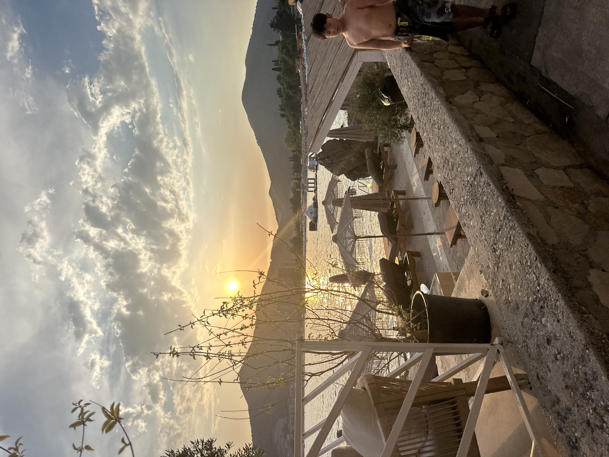 An outdoor, eye-level shot captures a young boy standing near a stone wall on a beach in Budva, Montenegro, during sunset. The boy, wearing dark, patterned shorts and no shirt, holds a yellow object, possibly a paddle, in his right hand. Behind him, a beach with loungers and umbrellas is visible, leading to the calm, shimmering sea. In the distance, lush green mountains rise, silhouetted against the golden glow of the setting sun, which casts a warm light on the scattered clouds. A white railing and a potted plant are in the foreground, framing the scene.