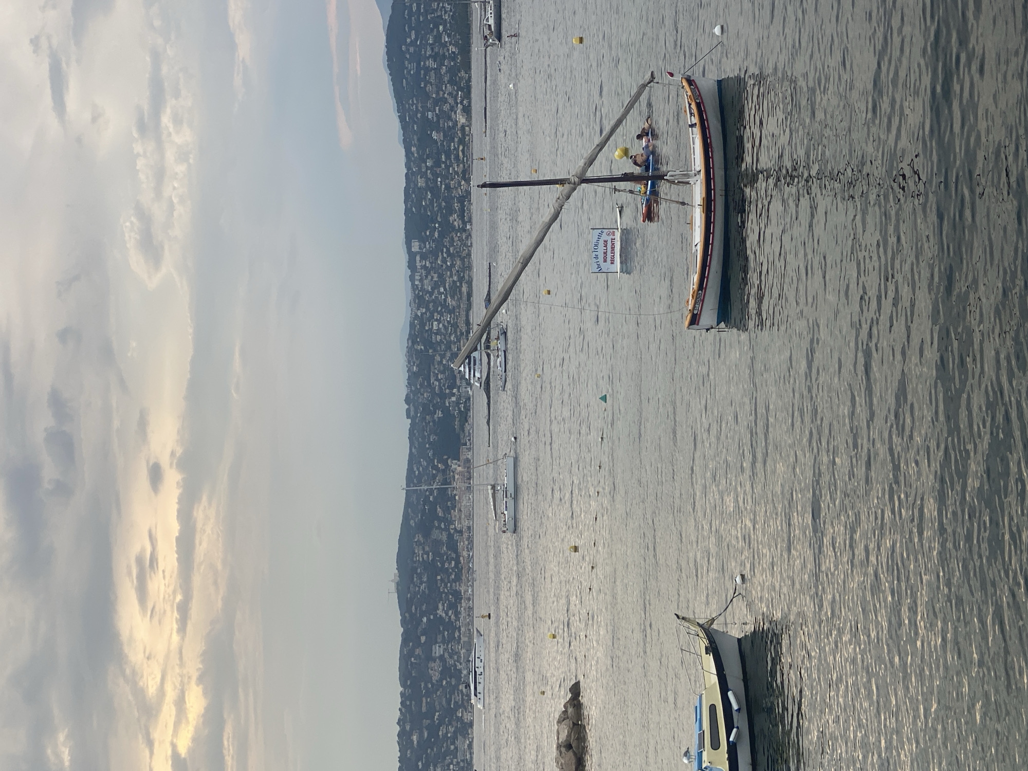 The image depicts a body of water, likely the Mediterranean Sea, with several boats and a coastal town in the background, consistent with the city of Antibes, France. In the foreground, a small, traditional wooden fishing boat with a single mast and sail is anchored. The sail is partially lowered and angled. There are people in the water near this boat, some appearing to be on a paddleboard. Another, larger, white sailboat is further in the distance, as is a white luxury yacht. The coastline behind the water is densely populated with buildings, characteristic of the urban landscape of Antibes. The sky is overcast with diffused light, suggesting either early morning or late afternoon. The water is calm, with gentle ripples reflecting the sky. Yellow buoys are scattered throughout the water.