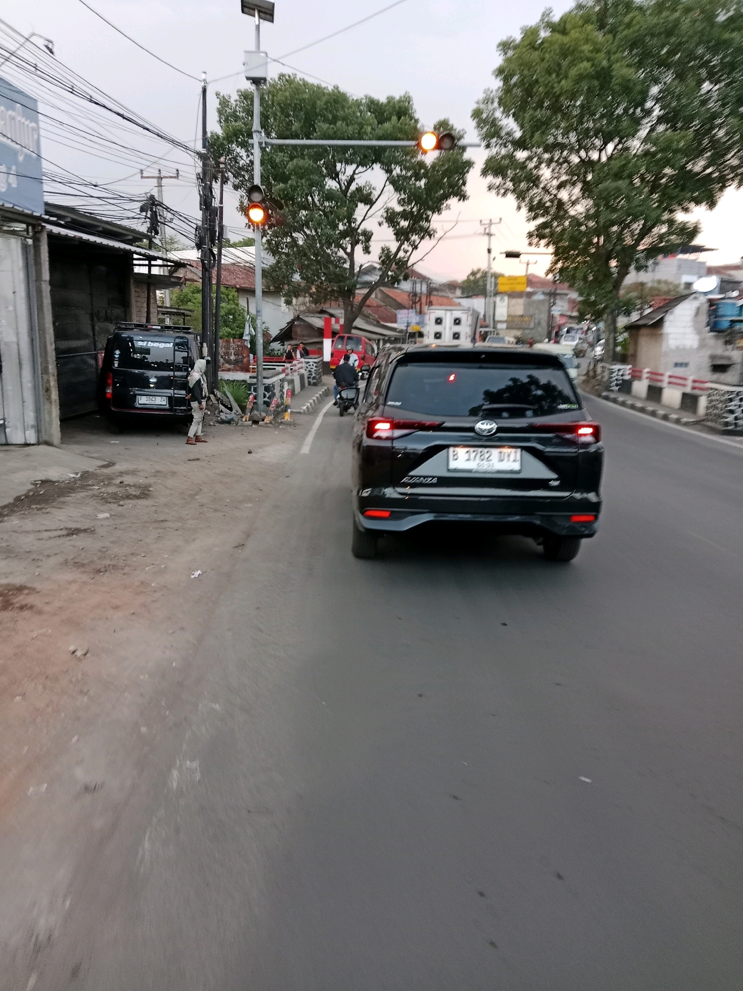 The image depicts a street scene in Sirnagalih, Indonesia, at what appears to be dusk. A black SUV, identified by its license plate "B 3782 DYI," is driving on the right side of the road. To the left of the road, a black van with the inscription "el begal" is parked. A person dressed in light-colored clothing and a headscarf stands on the sidewalk next to the van. Traffic lights are visible in the background, with one showing an orange light. The environment consists of a paved road, a sidewalk, buildings, trees, and numerous electrical wires overhead. The overall impression is of a moderately busy urban street.