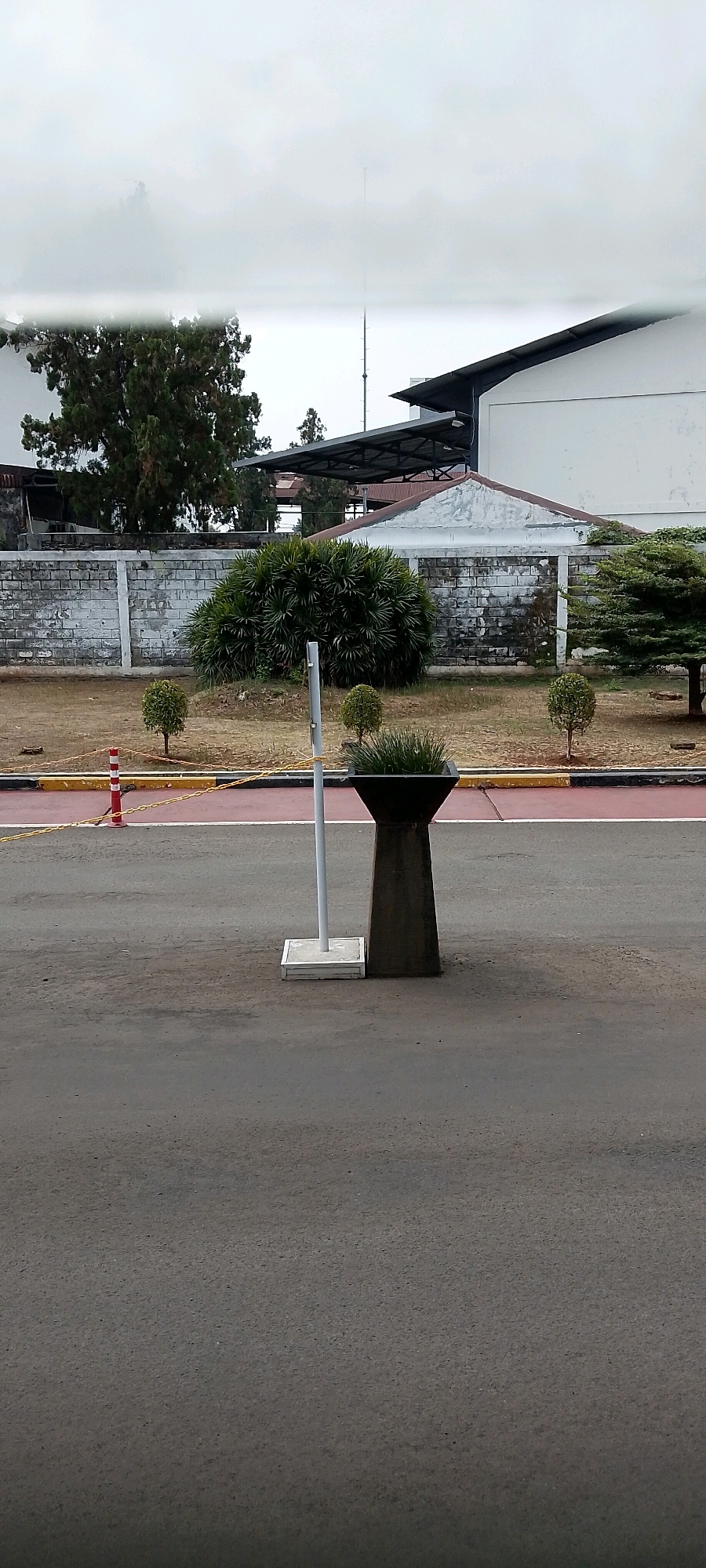 The image shows an outdoor scene with a road in the foreground and buildings and vegetation in the background. The road is asphalt and appears to be in good condition. There is a red and white striped traffic cone on the left side of the road, and a yellow chain barrier behind it. In the middle ground, there is a concrete planter with grass in it, and a white pole next to it. Beyond the planter, there is a low white brick wall, with a lush green bush in front of it. To the right of the bush, there are two small trees and a larger tree in the far distance. In the background, there are industrial-style buildings with white walls and dark roofs. The sky is overcast. The image is taken from a slightly elevated position, looking towards the buildings. The context of Depok, Indonesia, suggests a tropical or subtropical climate, which is consistent with the presence of lush vegetation. The overall scene depicts a secured or industrial area, possibly a campus or a facility, with road access and landscaping.